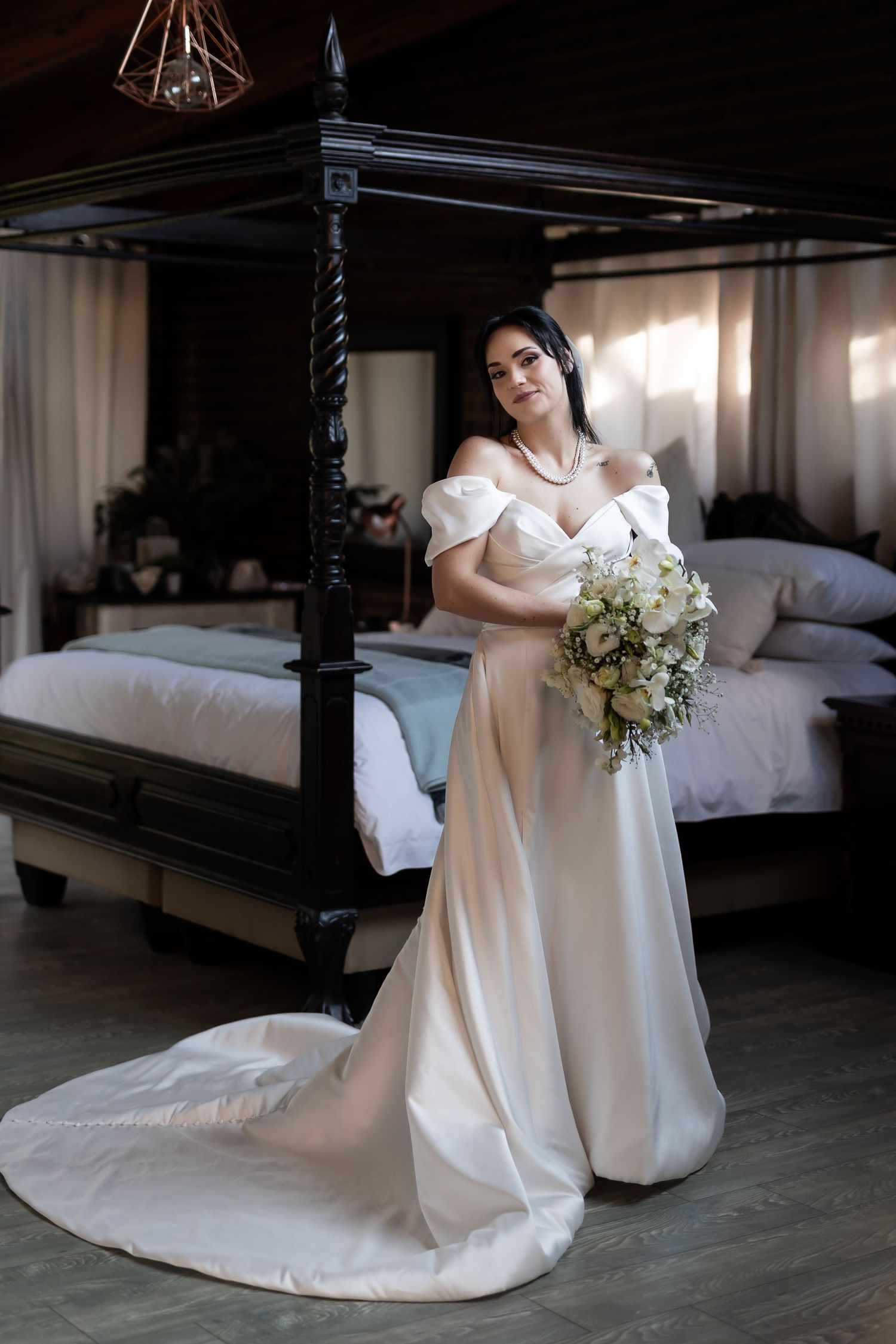 An elegant white wedding dress with off-shoulder design and flowing train is showcased in a bedroom with a dark canopy bed.