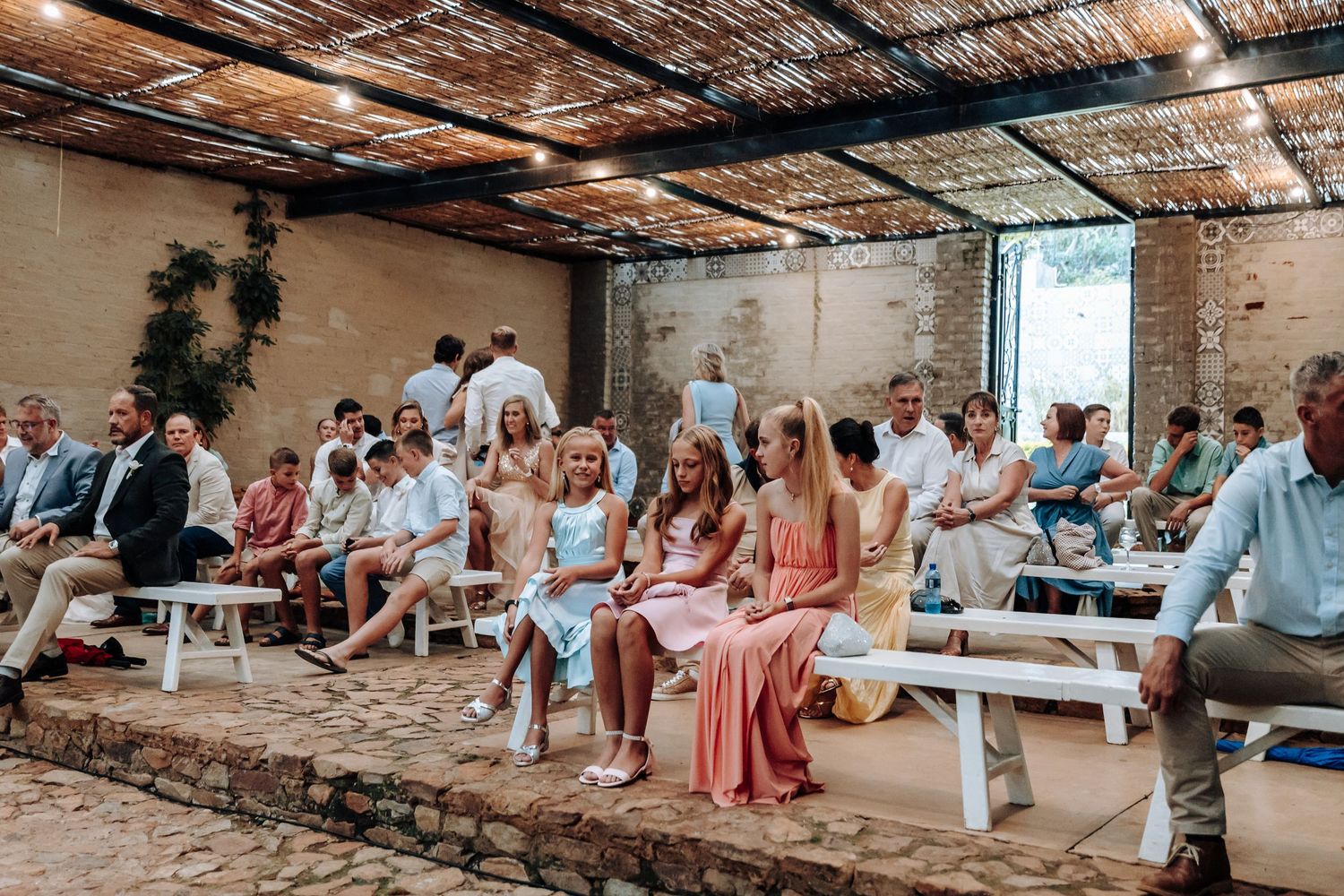 Guests seated on white benches at an outdoor rustic wedding ceremony under a pergola with stone walls.