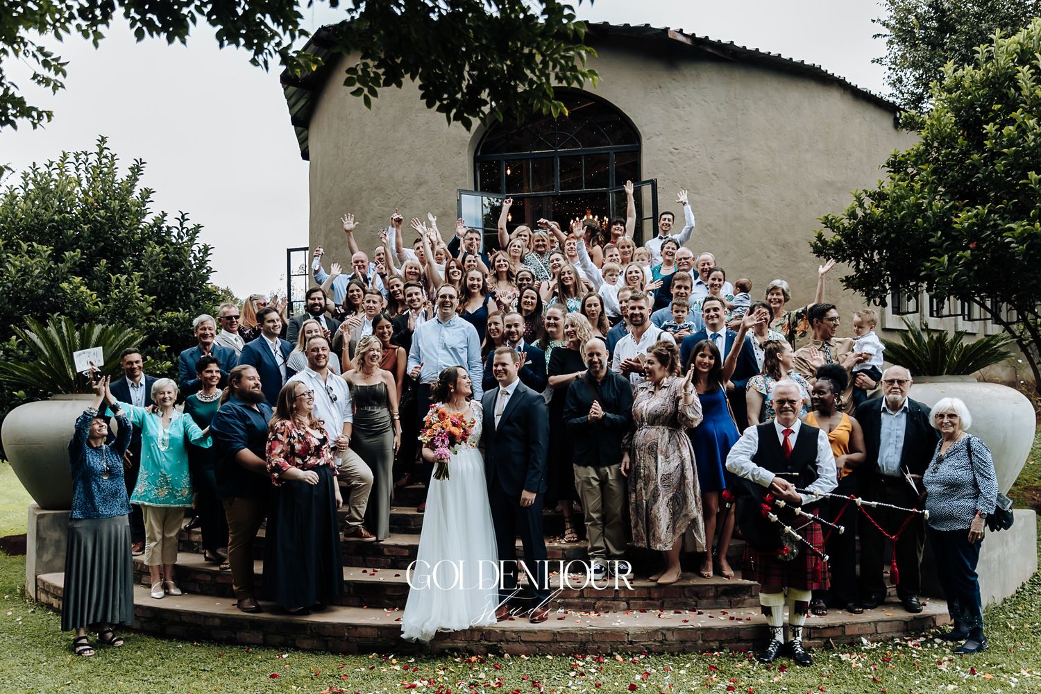Large wedding group photo with bride and groom surrounded by cheering guests outside venue.