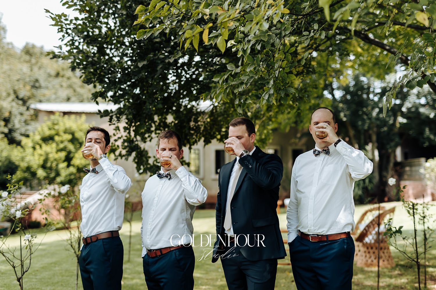 Groom and groomsmen in navy suits take a celebratory shot together outdoors at a wedding.