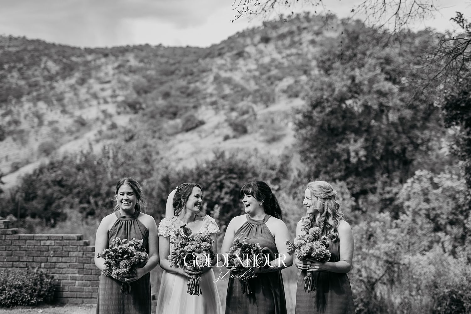 Bride and bridesmaids in dark dresses holding bouquets with lush mountain greenery in the background.