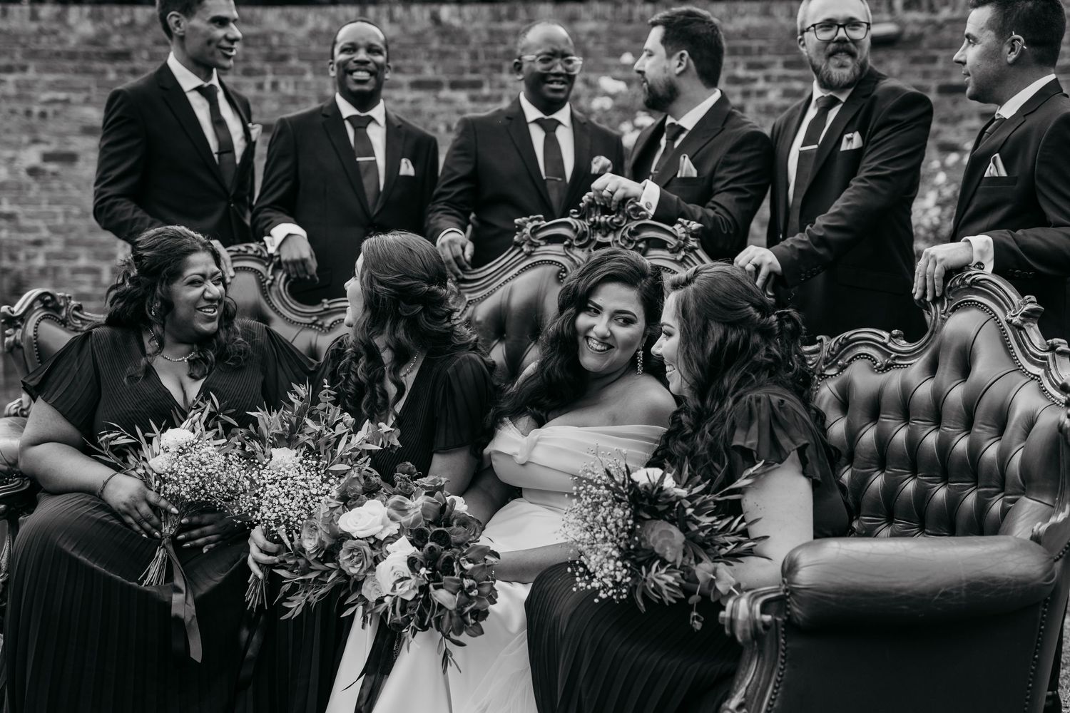 Wedding party shares candid laughs and moments on an elegant tufted sofa outdoors.