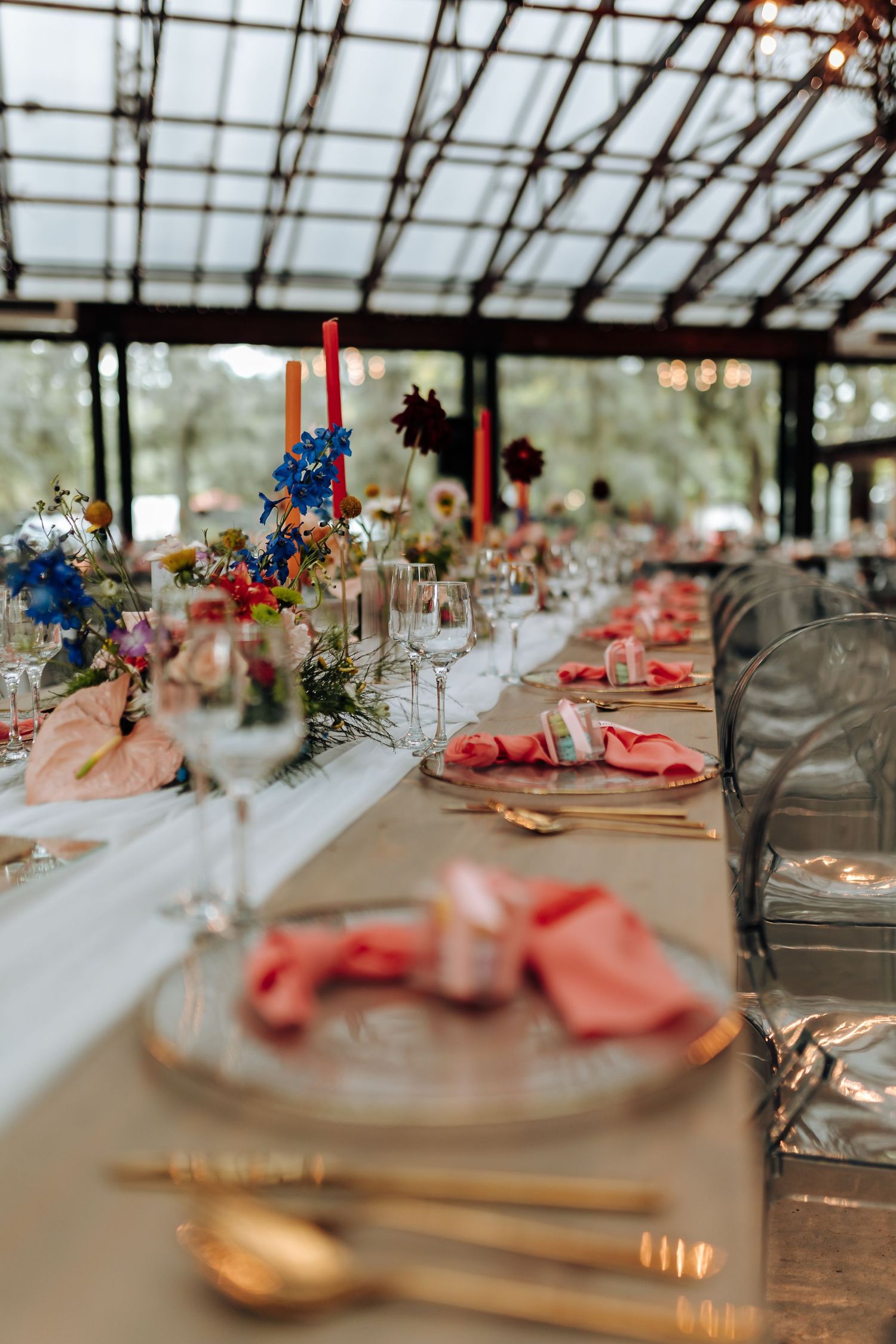 Long banquet tables set with coral napkins and blue floral centerpieces in an elegant greenhouse reception setting.