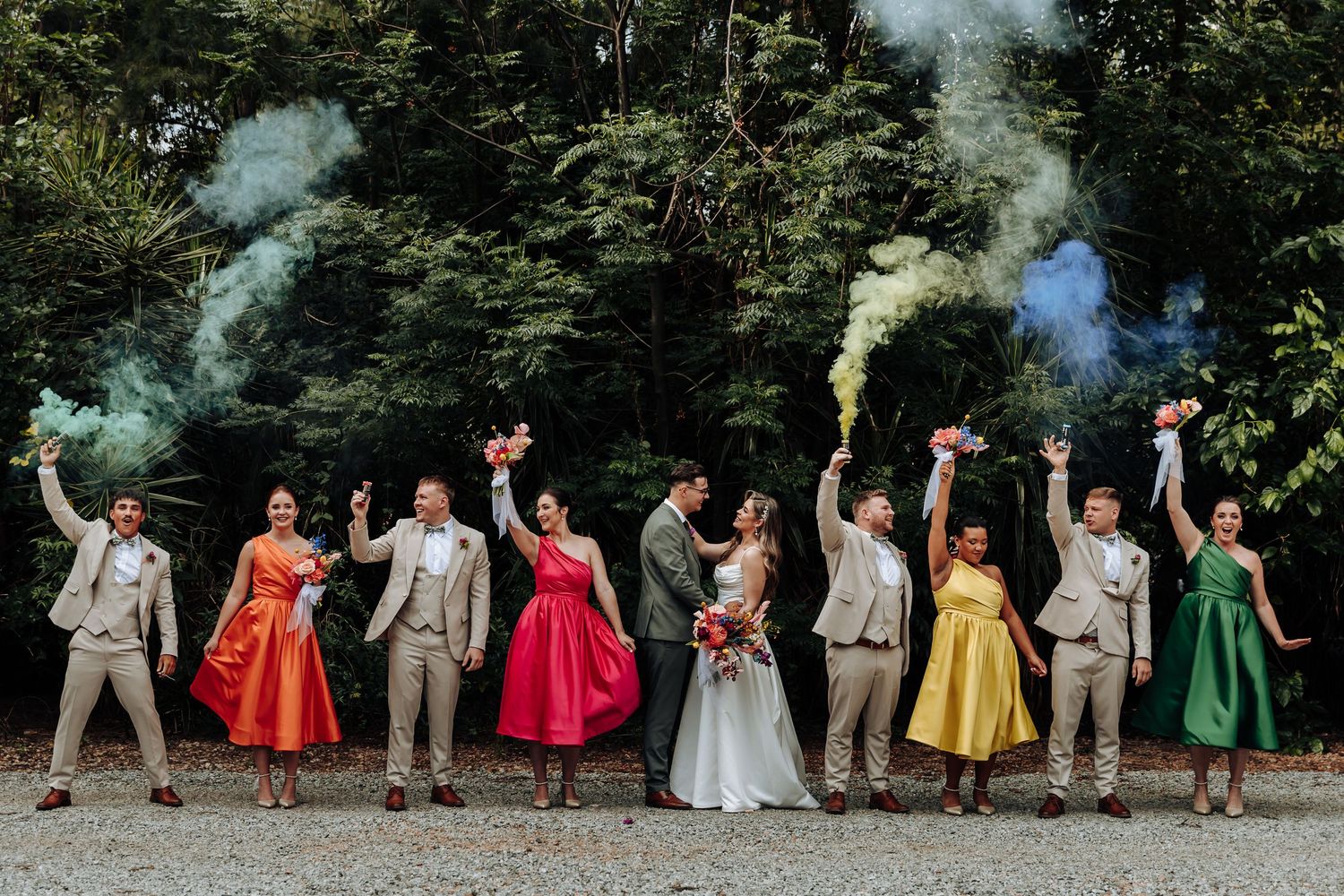 Wedding party in colorful dresses and grey suits poses together for a group photo in a forest setting.