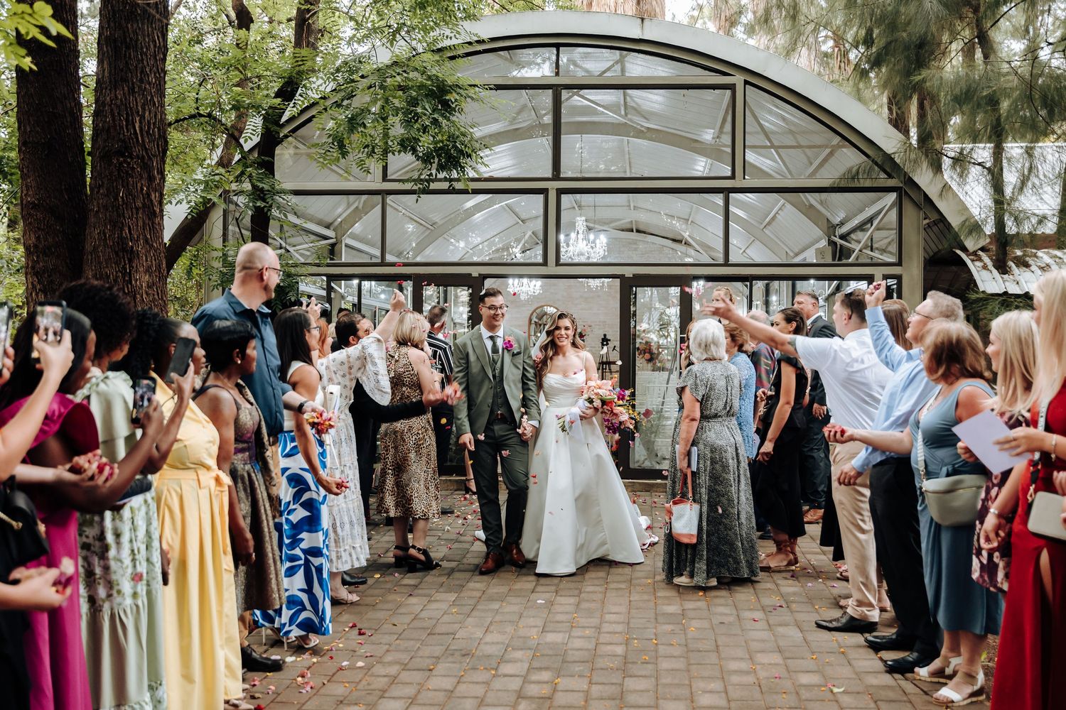 Wedding guests celebrate under an arched greenhouse structure during an outdoor garden ceremony.