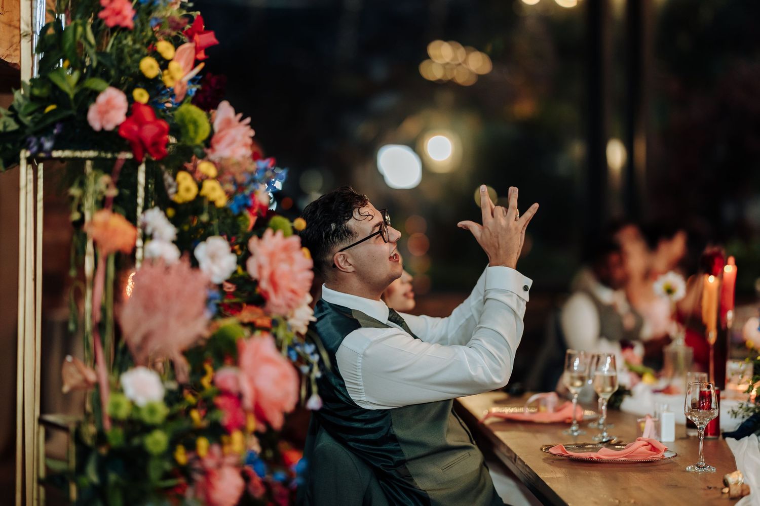 Wedding guests dancing and celebrating at reception with colorful floral decorations in background.