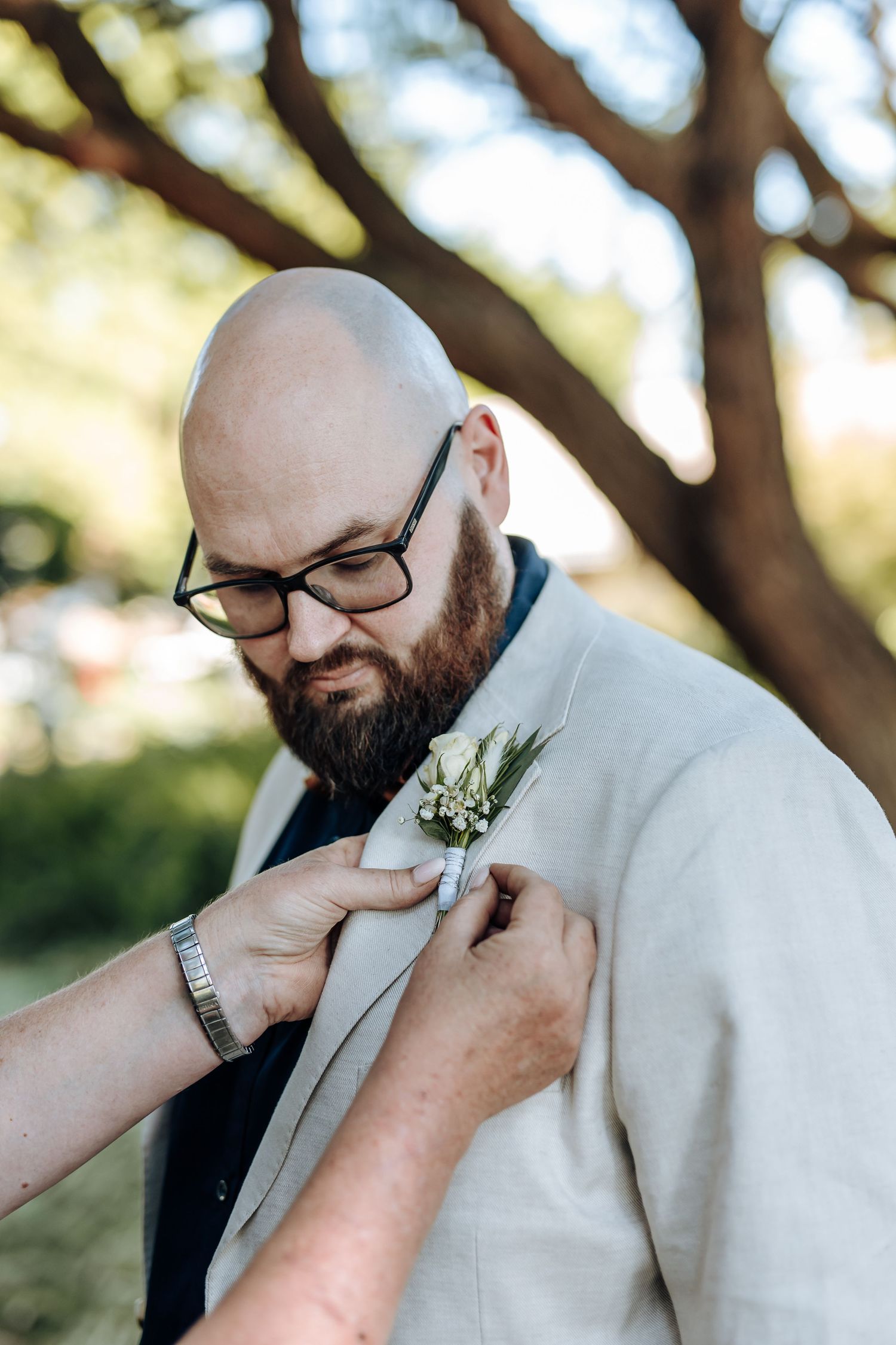 A person in a light gray suit jacket and glasses smells a small white boutonniere in a series of photos.