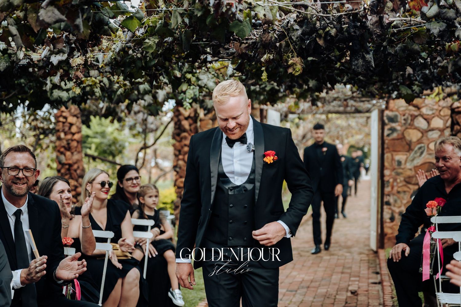 Wedding guests seated on chairs along a brick pathway in a garden setting with string musicians performing.