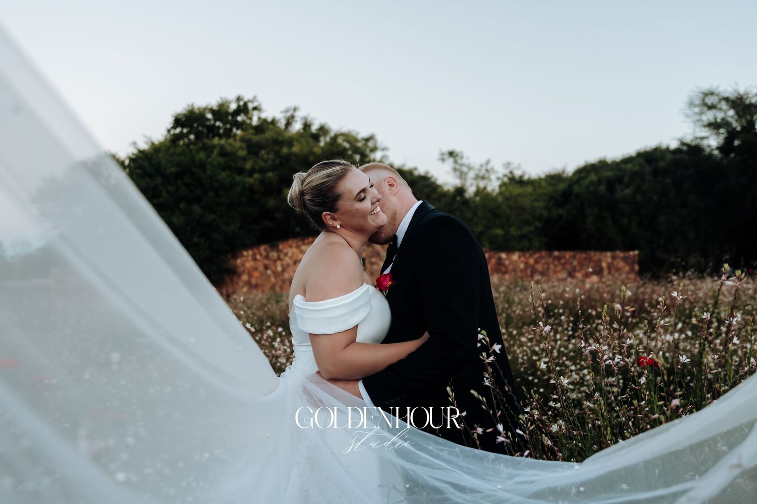 A romantic couple embraces in a field of white wildflowers as a long veil flows dramatically in the wind.
