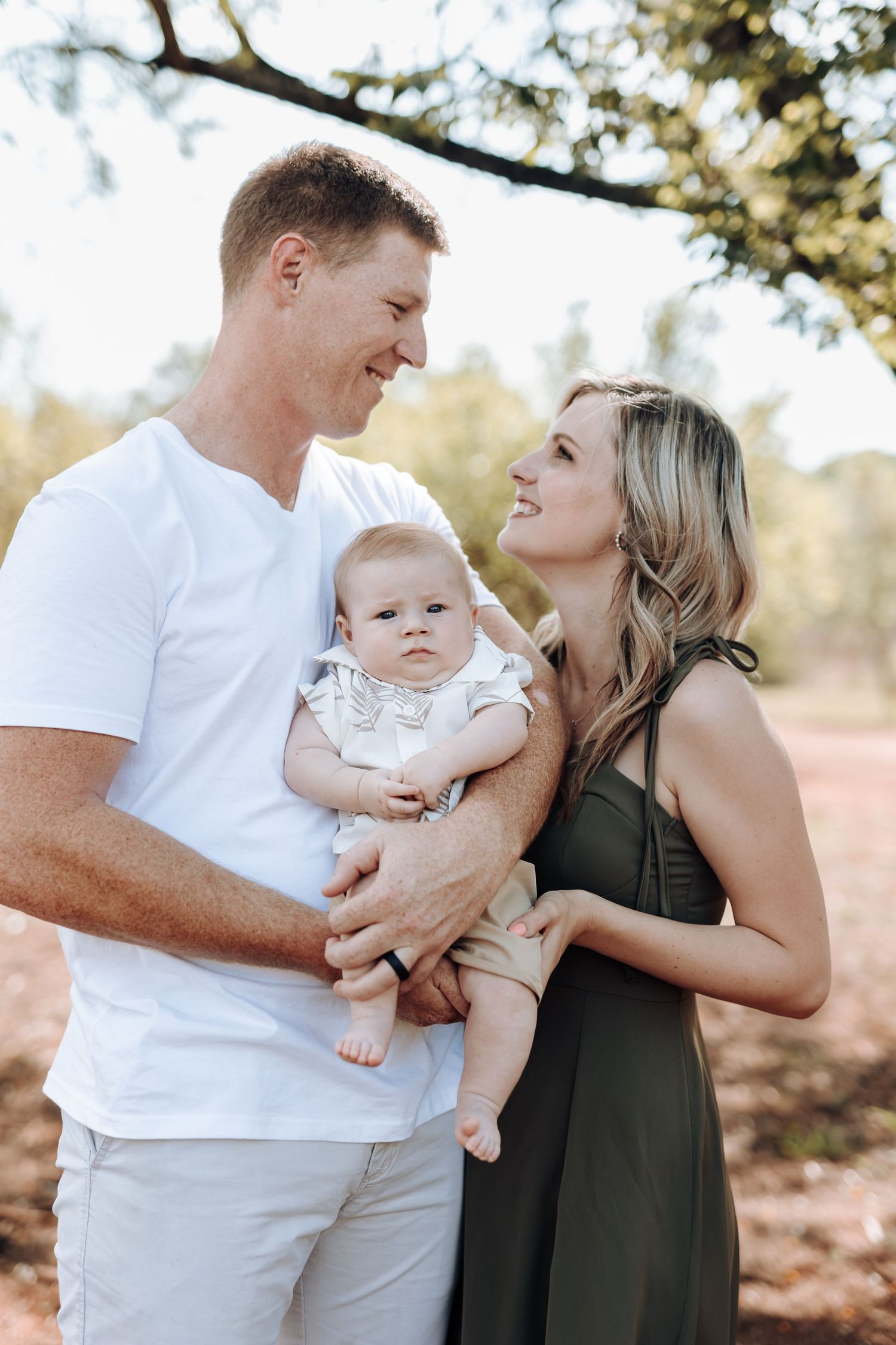A couple in white and olive green outfits share tender moments with their baby during an outdoor photoshoot.