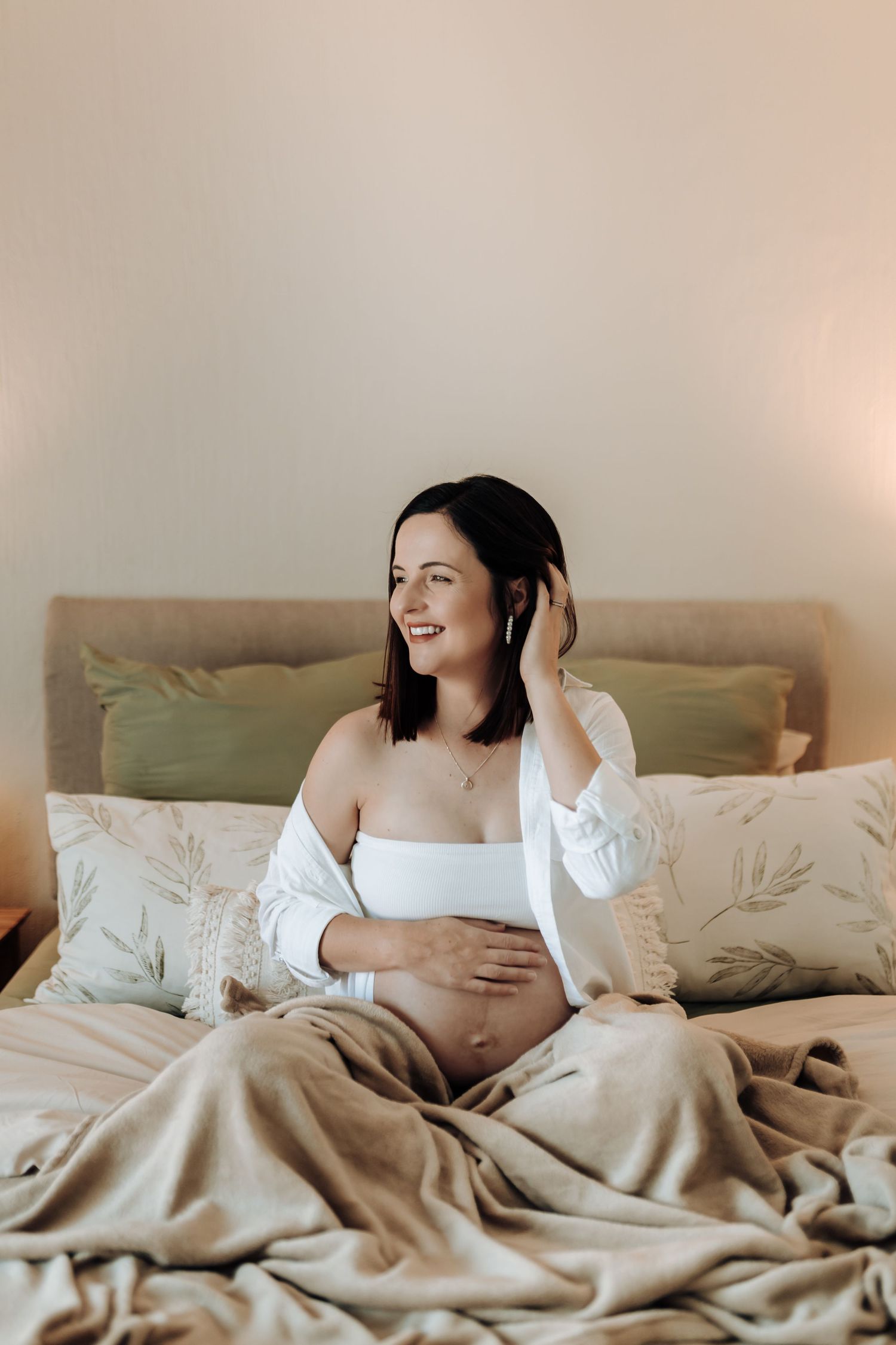Artistic maternity boudoir photos with subject reclining on bed wearing white shirt with beige bedding.