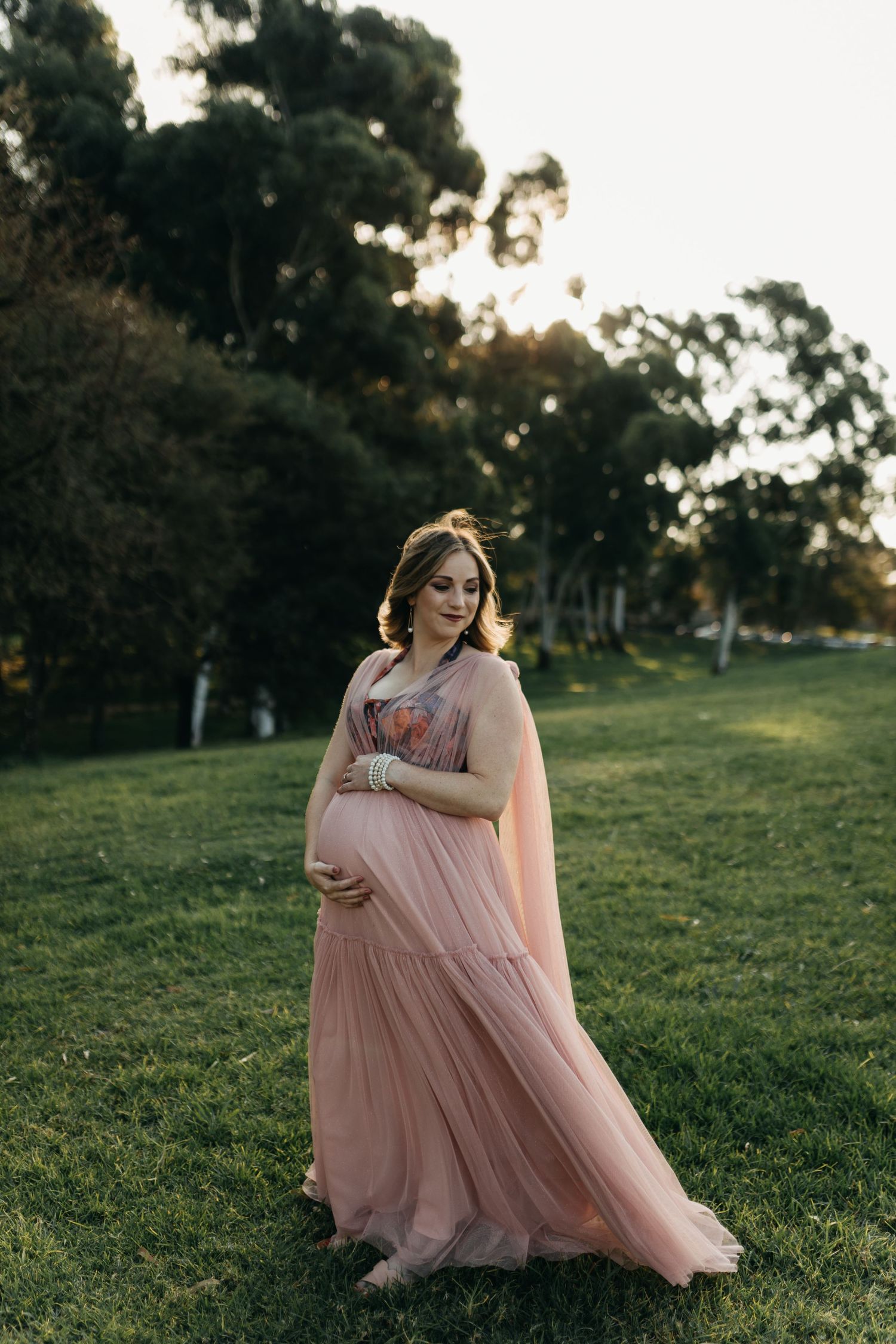 A person in a flowing blush pink dress stands gracefully in a grassy field at sunset.