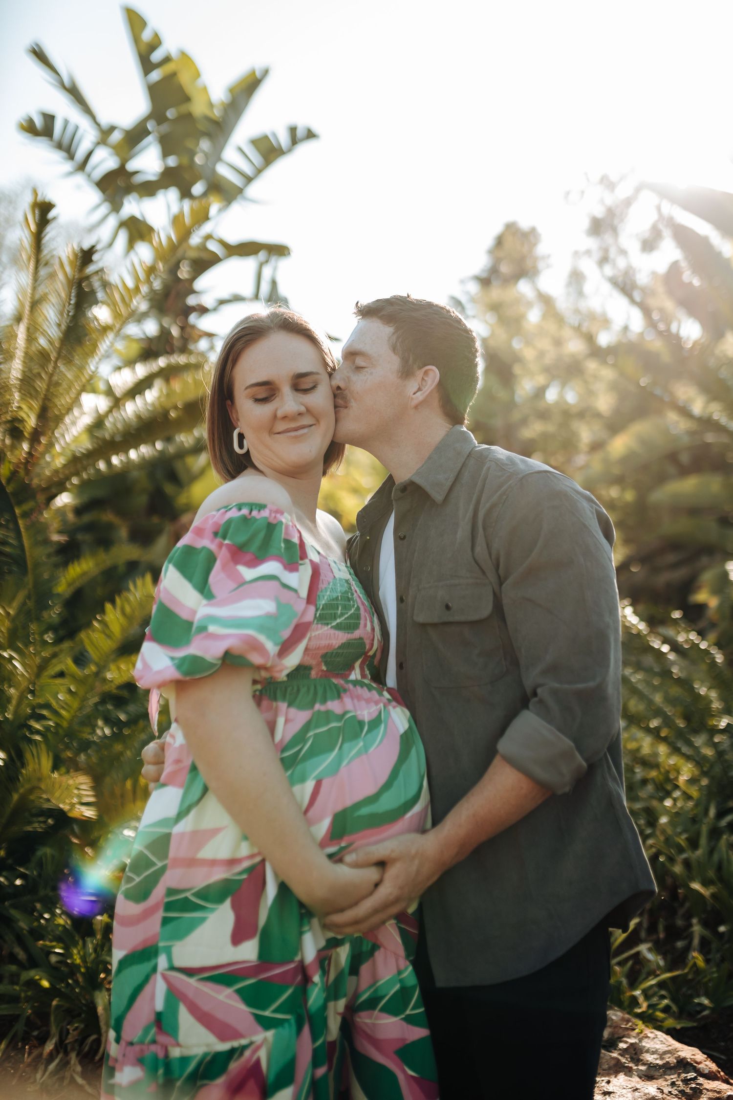 A romantic couple embraces in a tropical garden setting during golden hour sunlight with palm fronds in the background.
