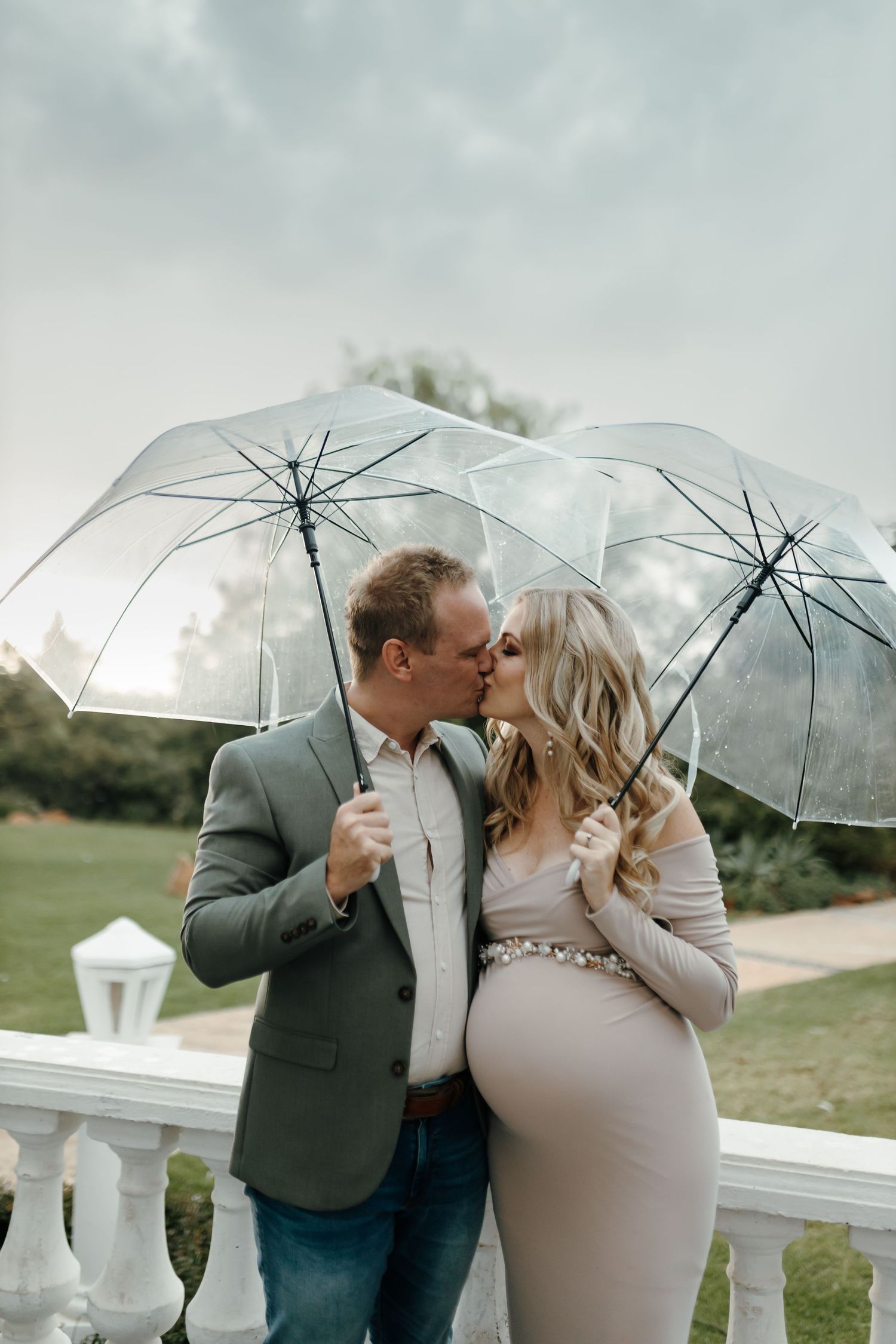 A couple poses with a clear umbrella during a romantic maternity photography session outdoors.