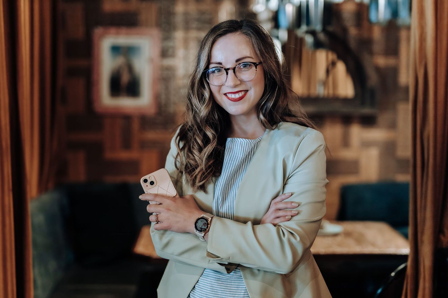 A stylish woman in business attire and glasses sits at a desk with a laptop in a warm-lit office space.