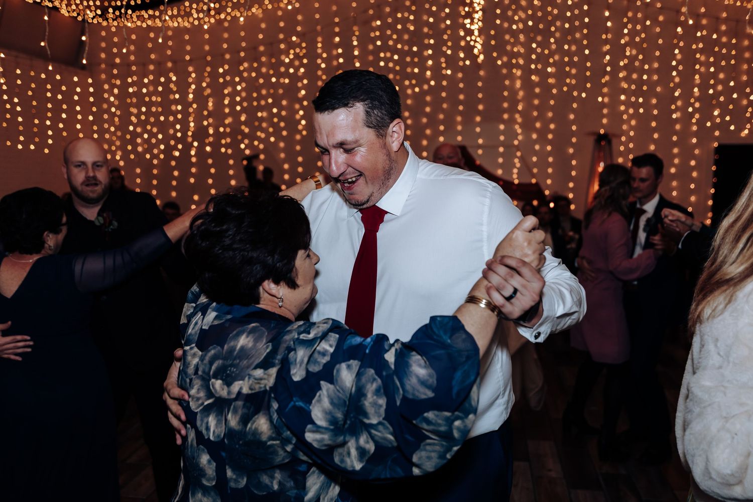 Guests dancing joyfully at a wedding reception with warm fairy lights glowing in the background.