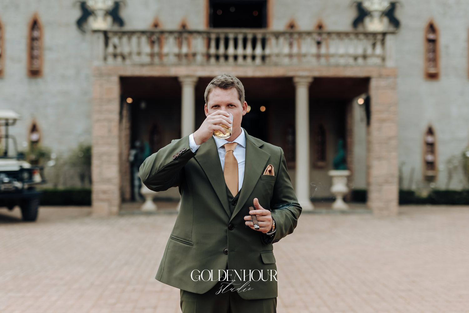 Groom in olive green suit drinking outside an elegant venue during wedding celebrations.