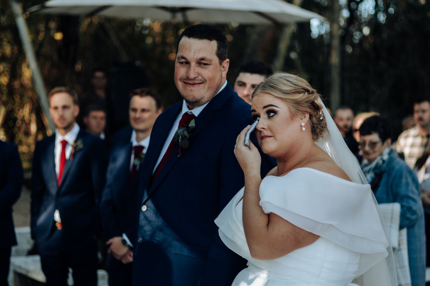 Emotional bride wiping tears beside smiling groom in navy suit during outdoor wedding ceremony.