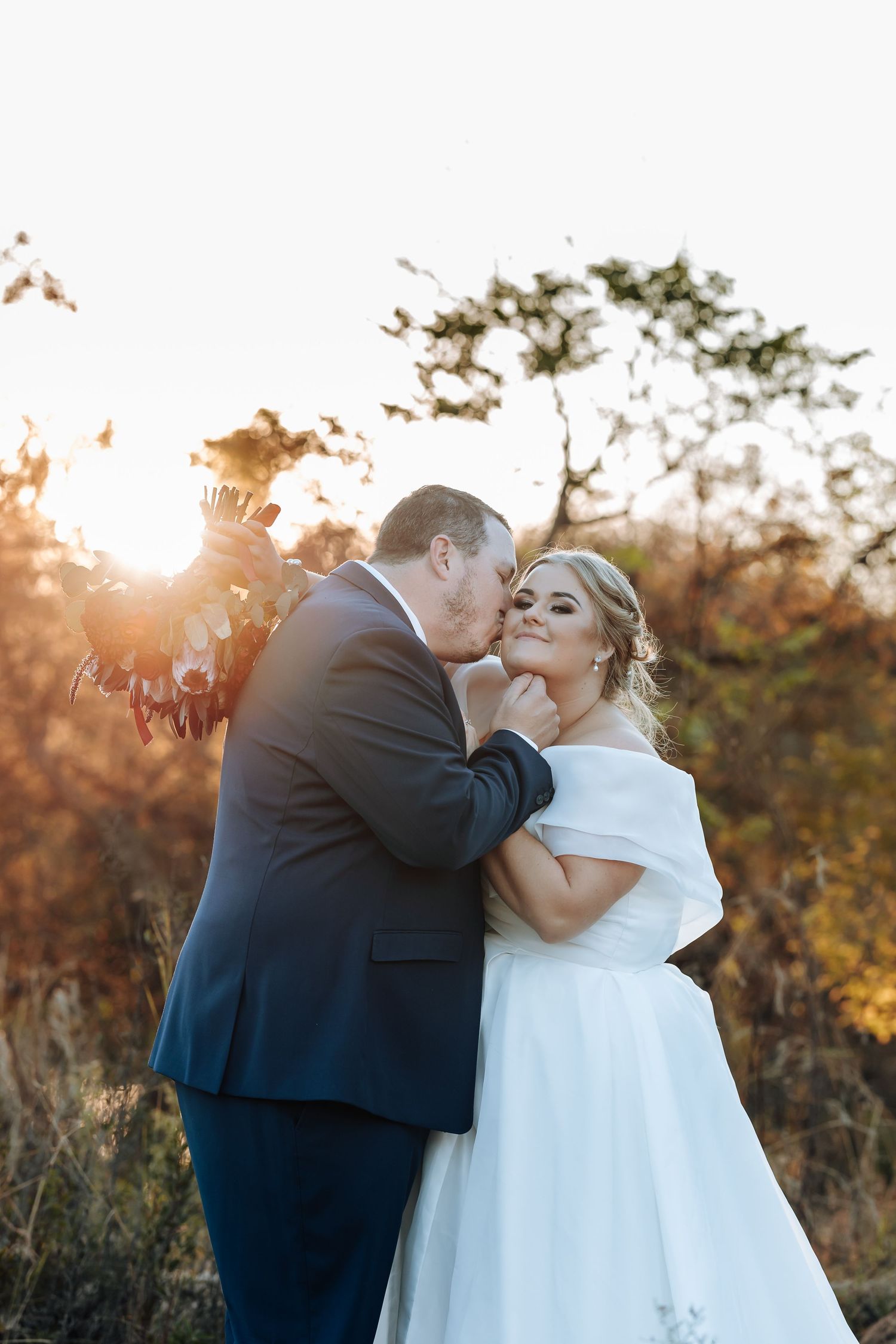 A groom in a navy suit kisses his bride in a white off-shoulder gown during a golden sunset outdoor wedding.