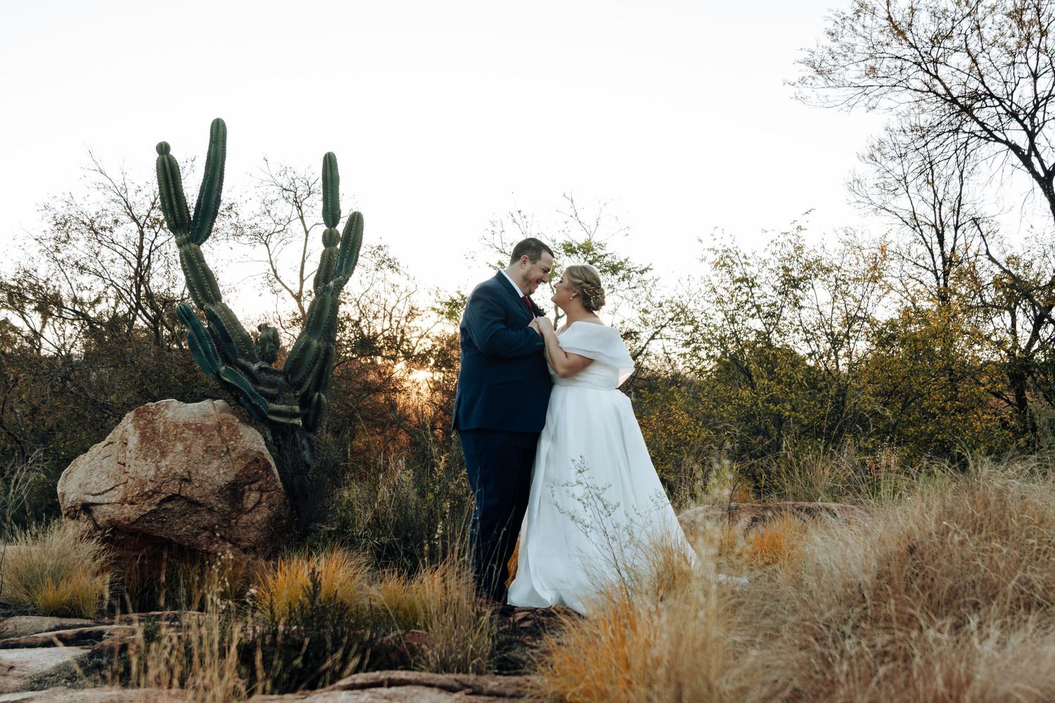 Bride and groom share romantic moment in desert landscape with saguaro cactus at sunset.