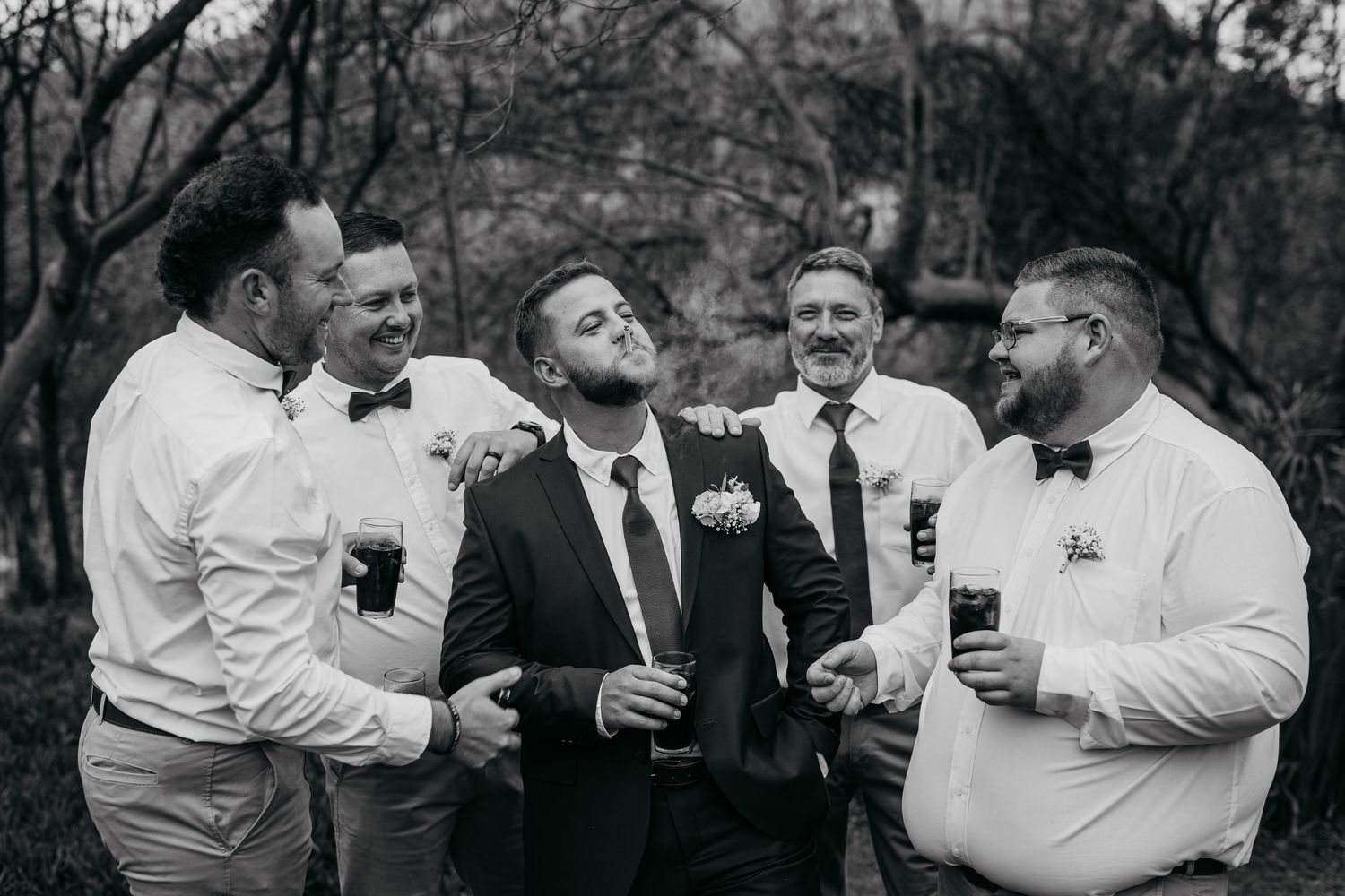 A group of men in formal attire laugh and embrace while holding drinks at what appears to be a wedding celebration.