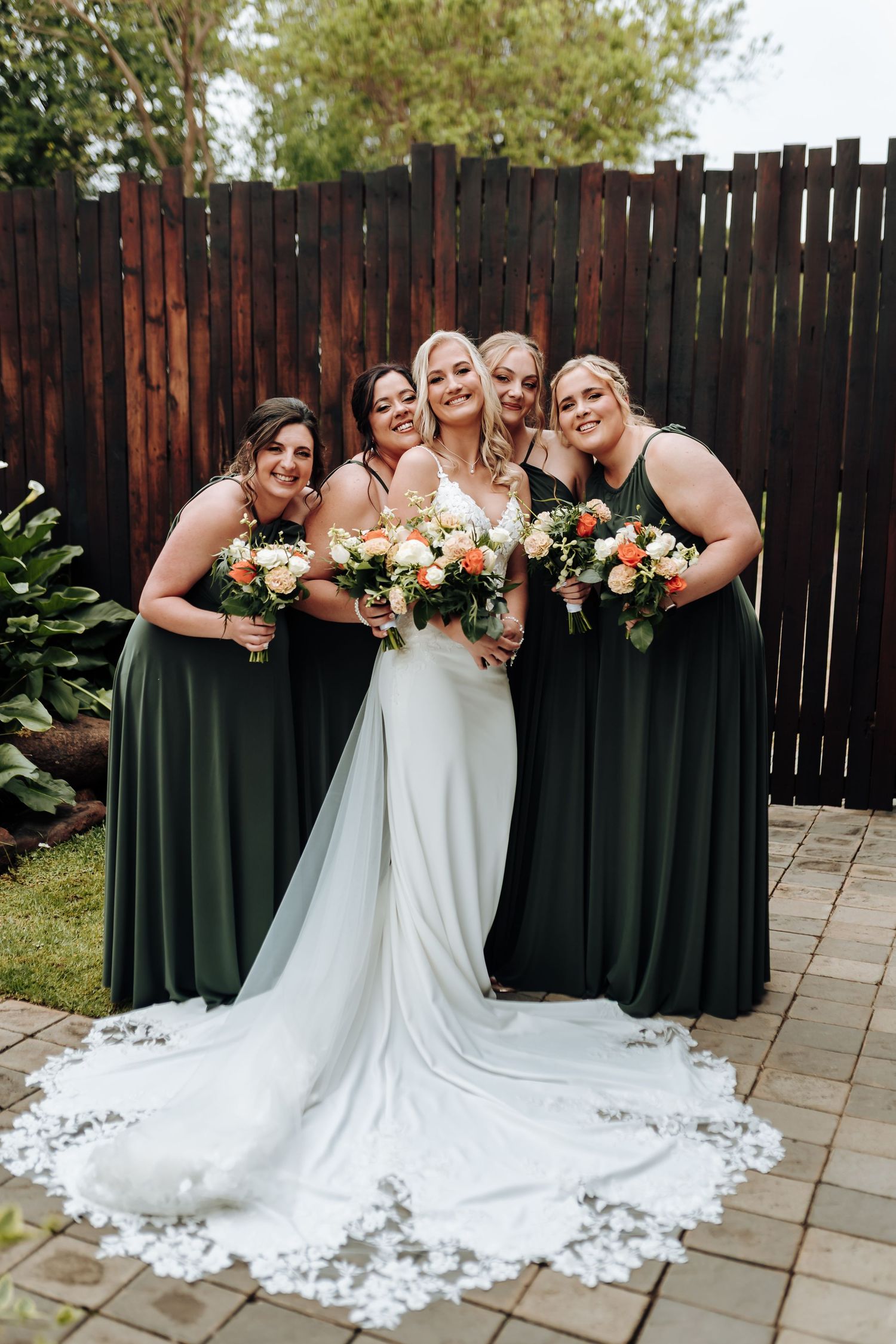 Bridesmaids in elegant dark green dresses pose with bride in white lace gown with long train at outdoor wedding venue.