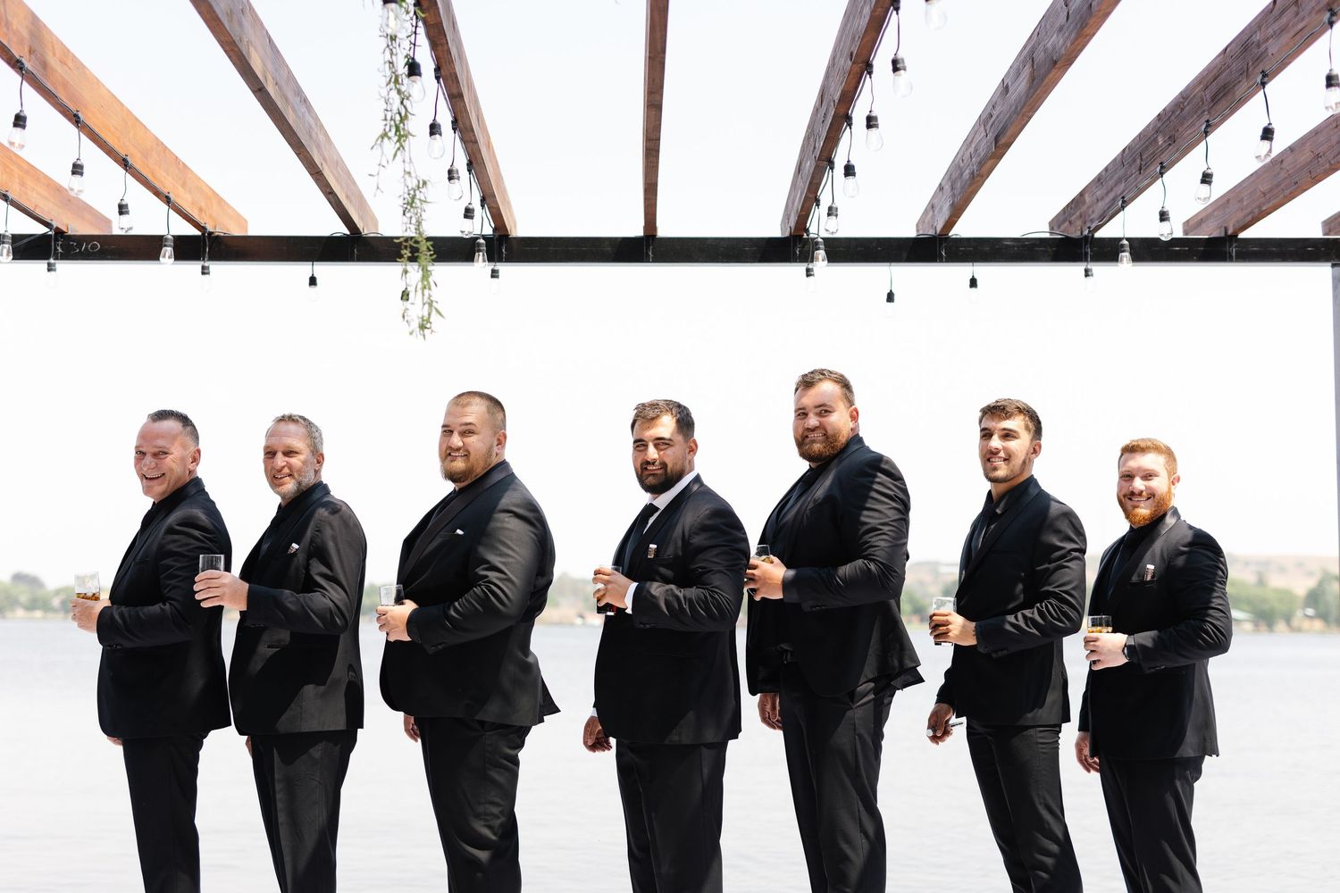 Groomsmen in black suits pose in sequence showing playful dance moves against a light colored background.