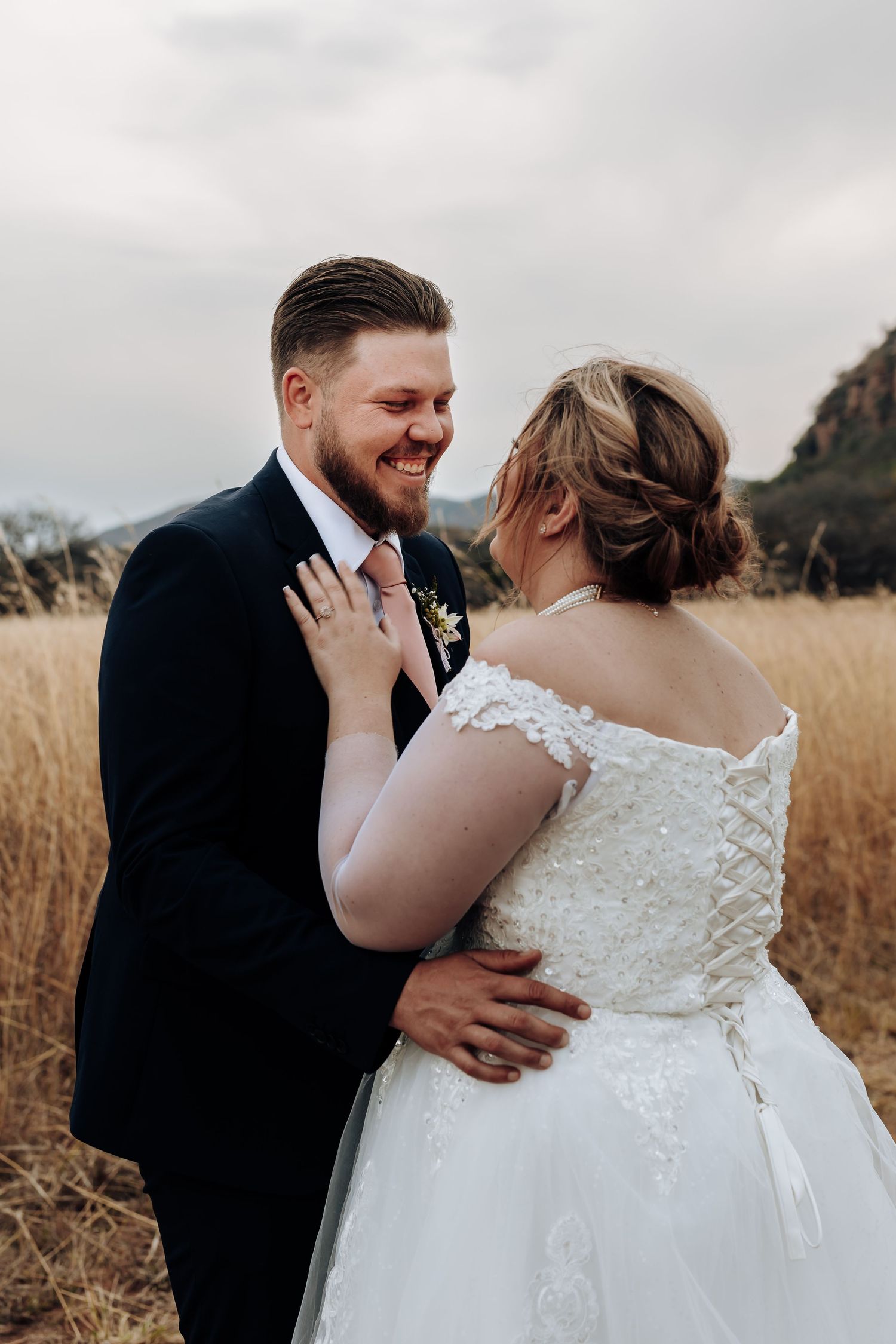 Newlywed couple share intimate moment in golden field during sunset with mountains in background.