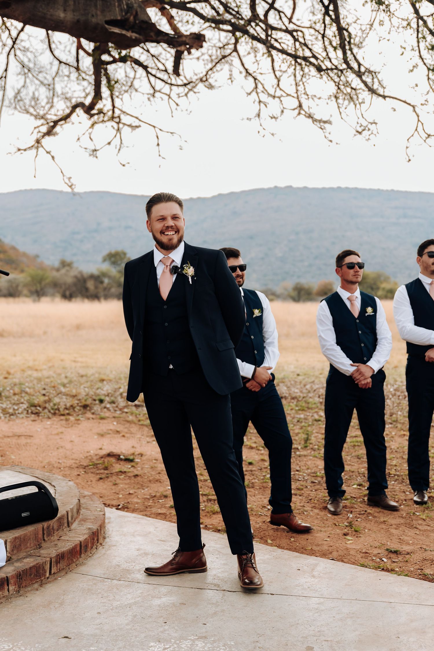 Wedding party members in black and white attire sit casually in a line outdoors with mountain scenery in background.