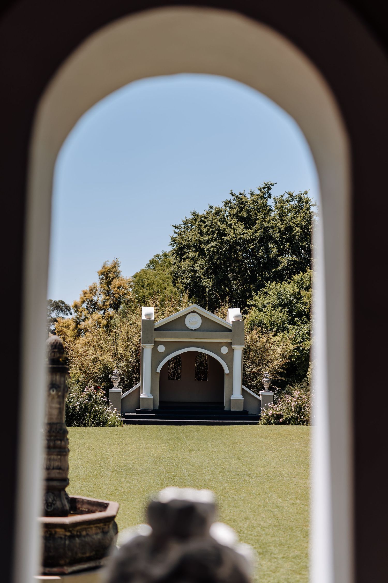 Small white chapel with arched entrance viewed through an archway frame on a sunny day with green lawn and trees.