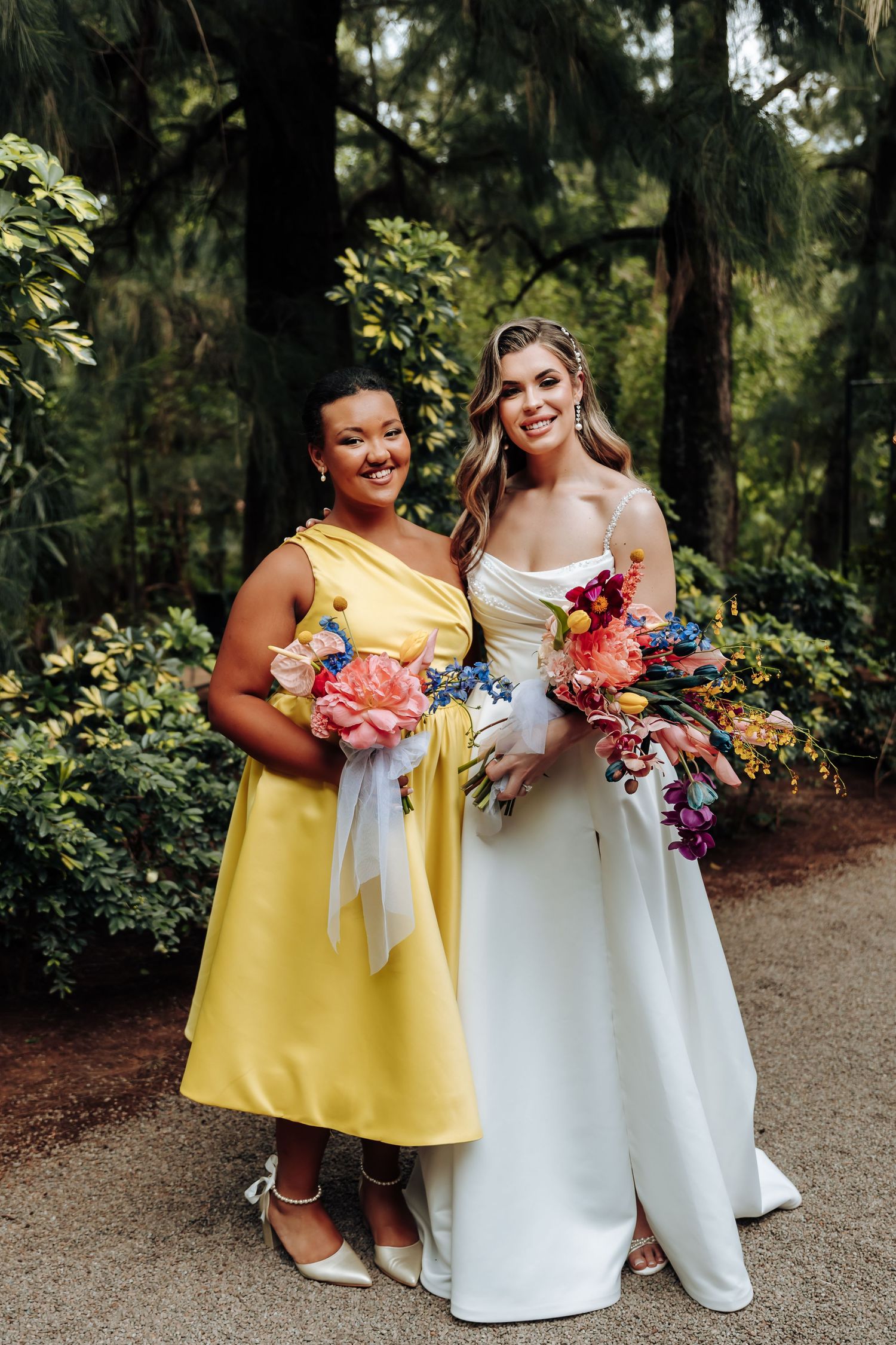 Wedding party members in yellow and white dresses hold colorful bouquets while posing together in a garden setting.