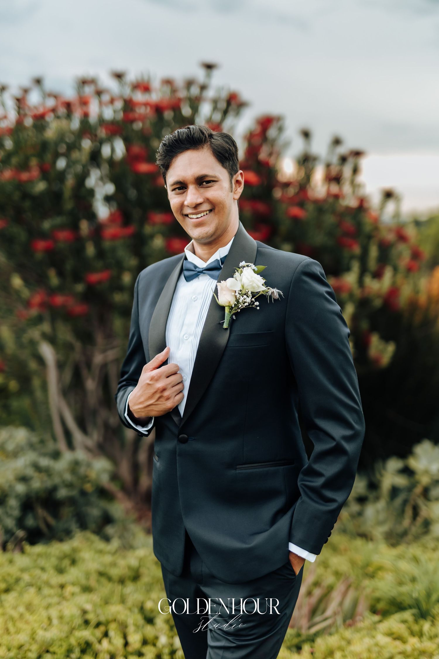 Groom in classic black tuxedo poses against flowering red bushes.