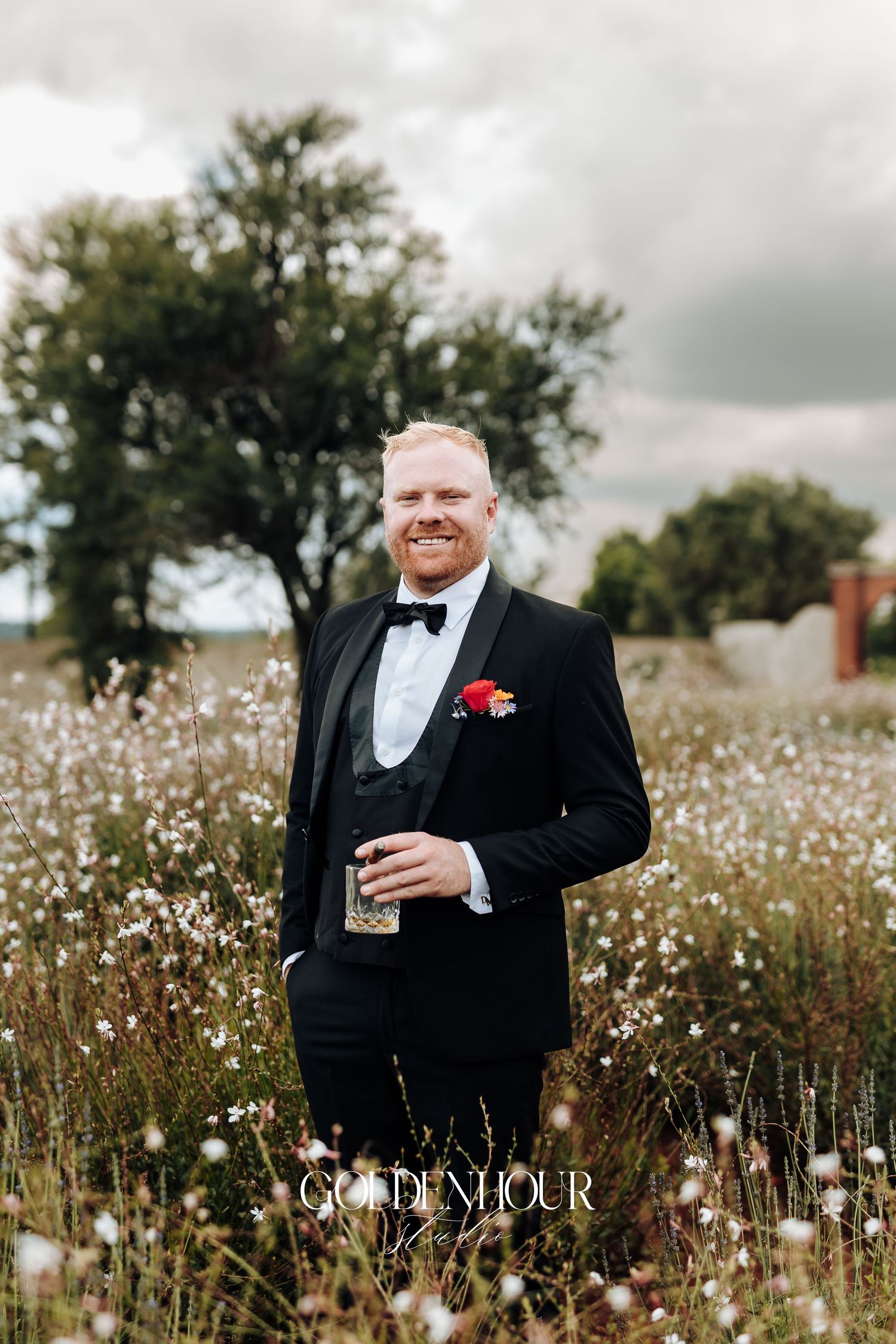 A person in a black tuxedo stands in a field of white wildflowers on a cloudy day.