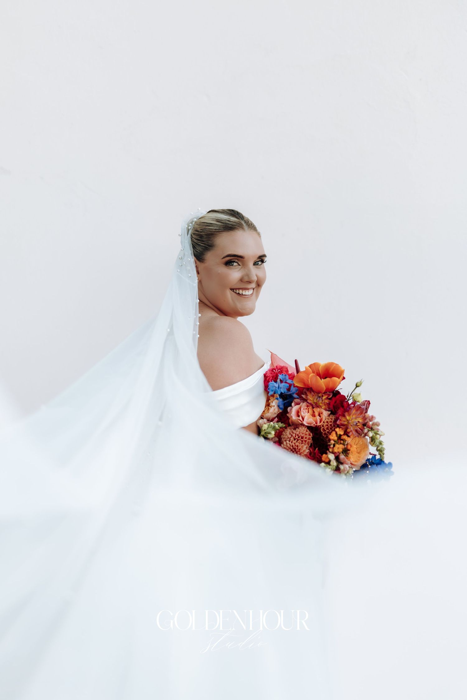 A bride in a flowing white veil holds a vibrant autumn-colored bouquet against a light background.