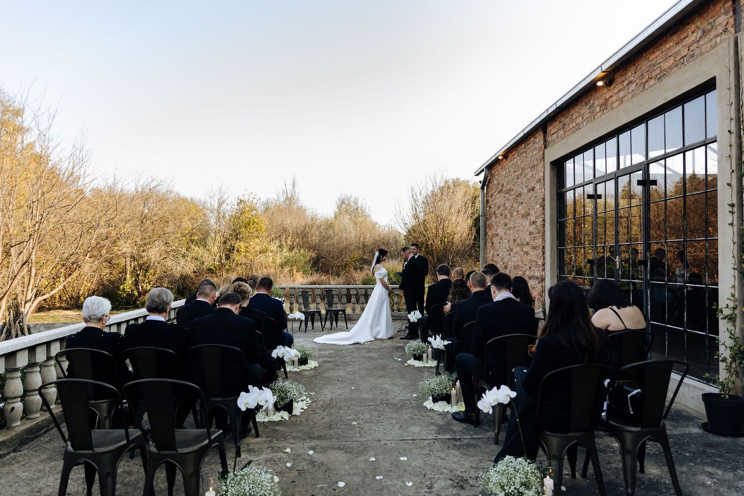 An outdoor autumn wedding ceremony setup with black chairs and stone walls along a terrace overlooking natural scenery.