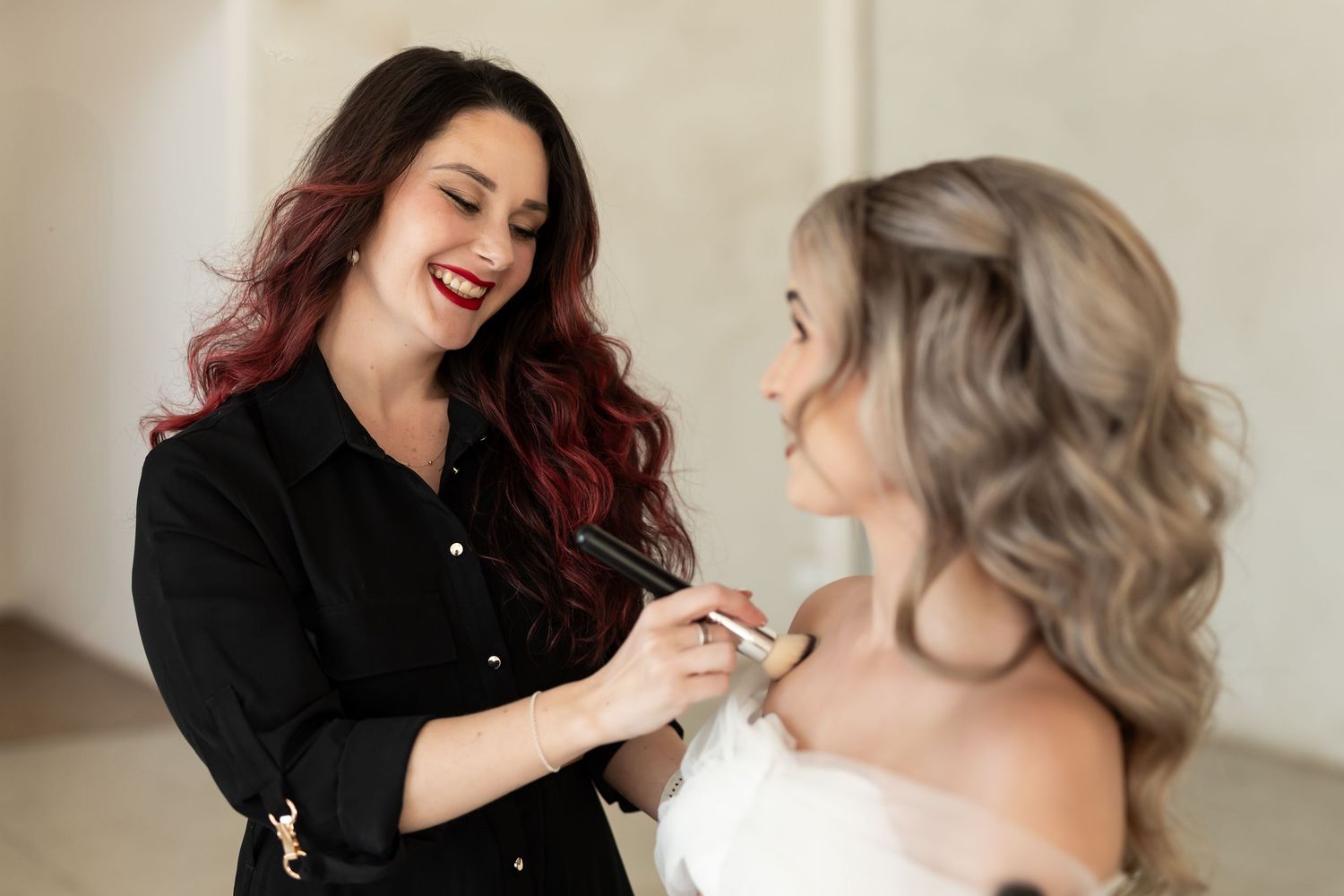 Professional stylist applies makeup in bright studio setting while client smiles during beauty session.