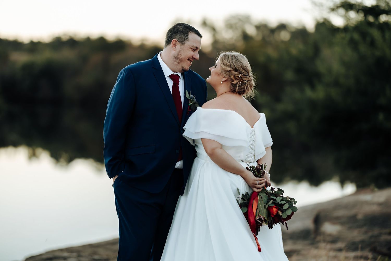 Bride and groom share a romantic moment by a lake, she holds a bouquet in a white off-shoulder gown, he wears a navy suit.