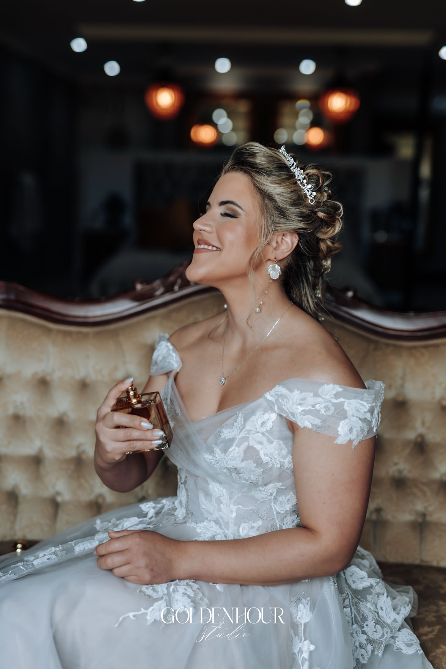 Radiant bride in lace off-shoulder gown and tiara sits smiling while holding perfume bottle.