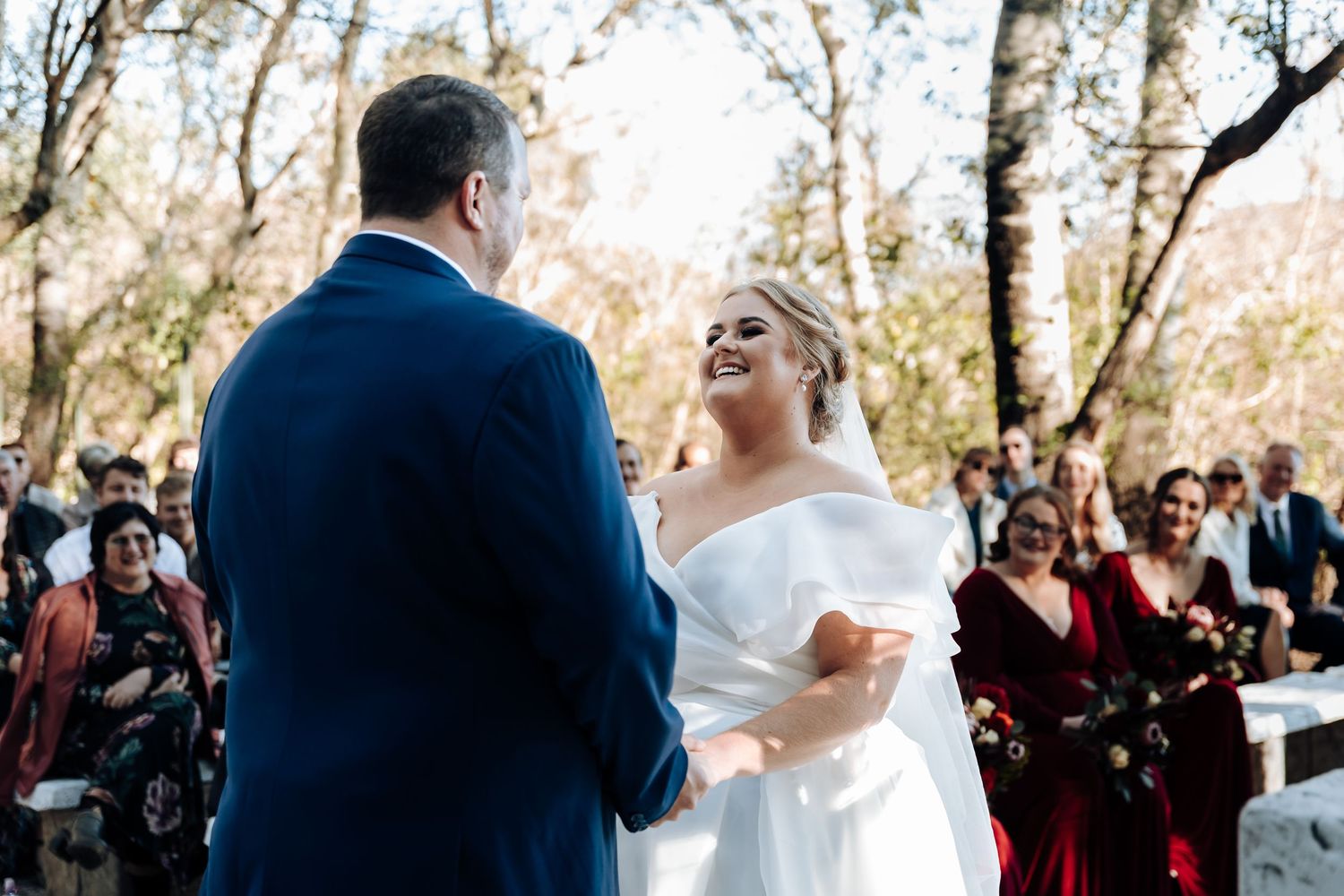 Bride smiling joyfully at groom during outdoor wedding ceremony with guests in background.