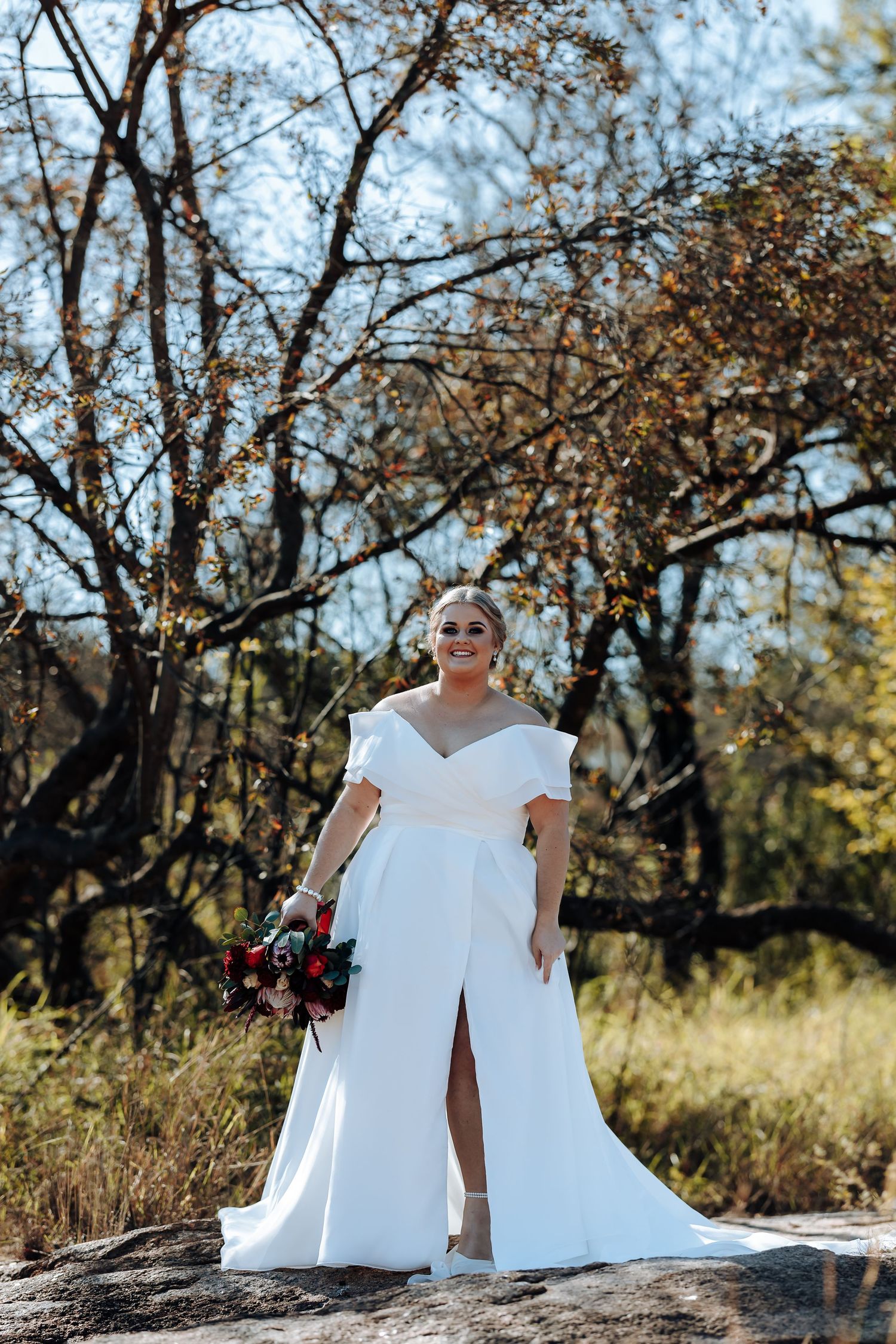 Bride in white off-shoulder gown with slit holds bouquet outdoors among autumn trees on rocky terrain.