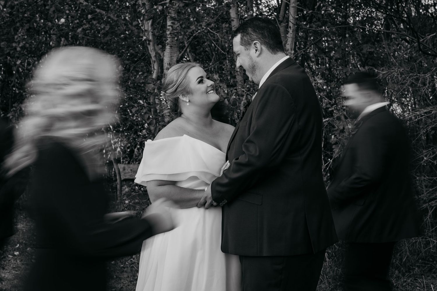 Black and white wedding photo of bride and groom sharing a tender moment outdoors, with blurred guests passing in foreground.