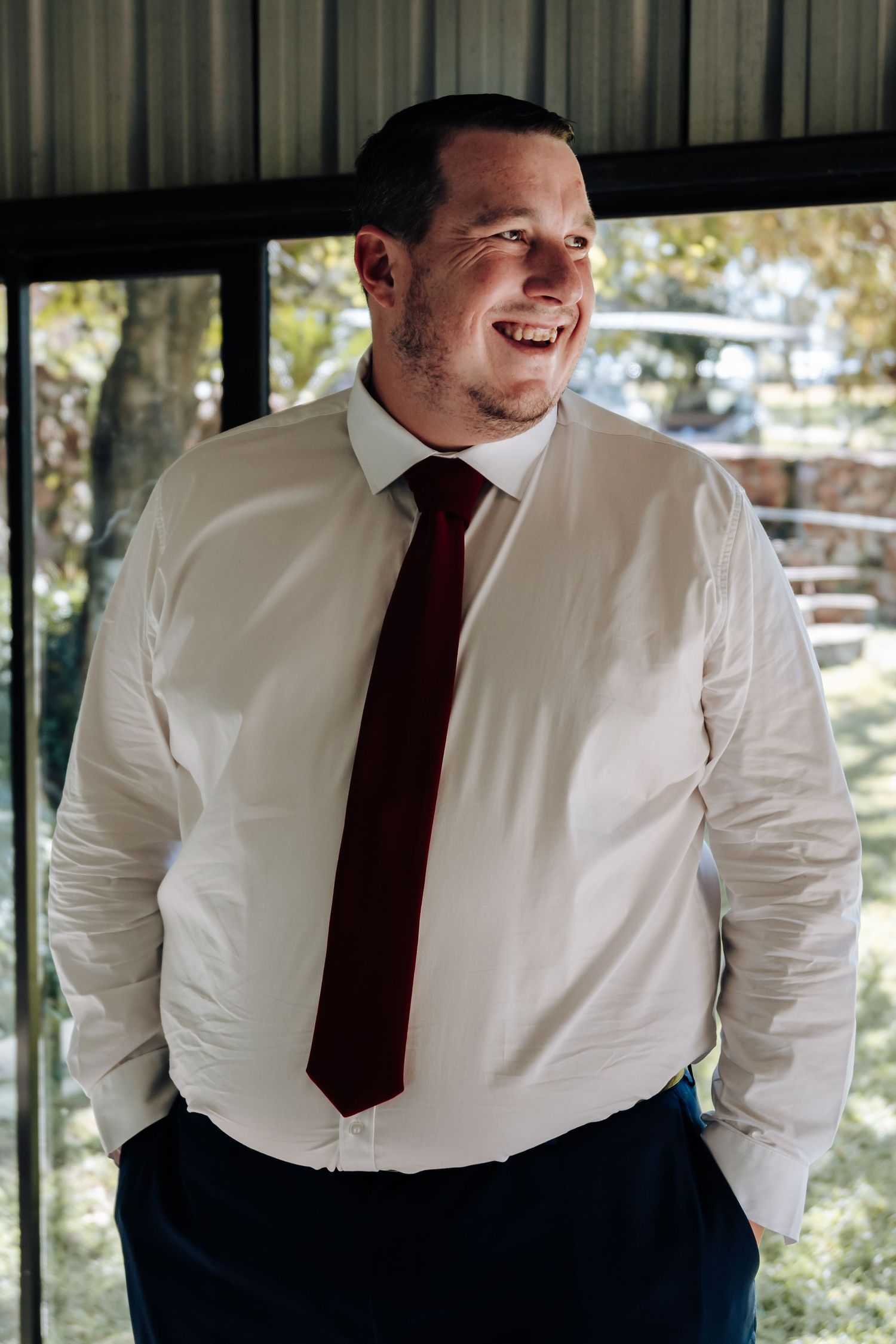 Smiling man in white dress shirt and burgundy tie standing indoors with hands in pockets.
