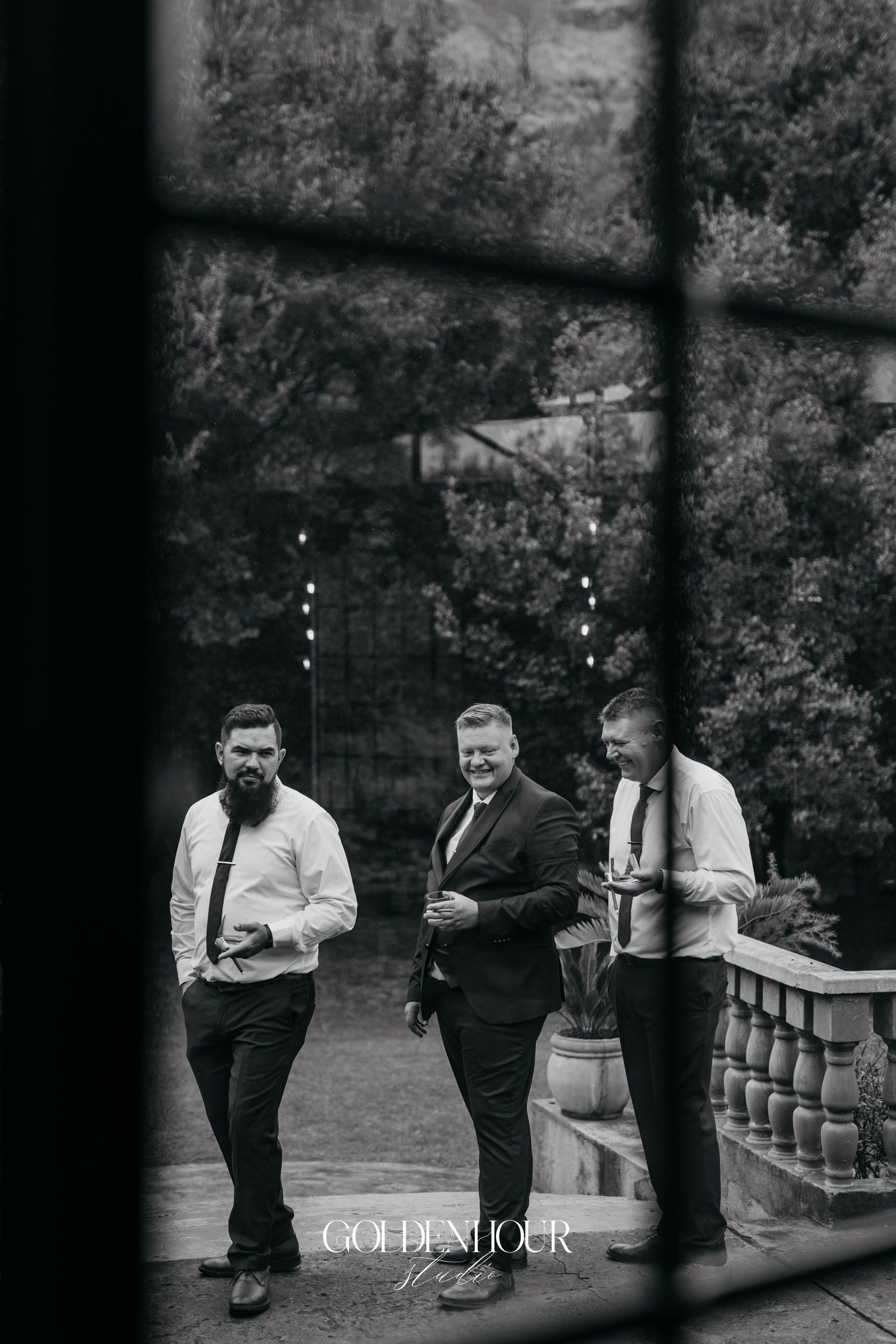 Black and white photo of three groomsmen walking on an outdoor terrace surrounded by lush greenery.