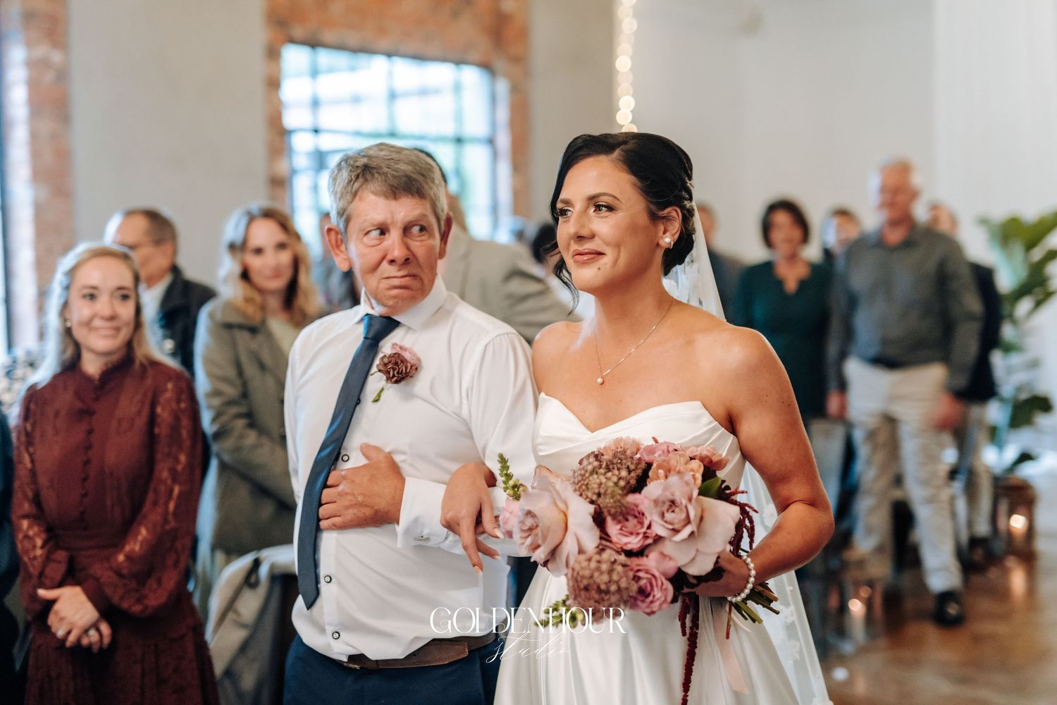 Bride walks down the aisle with her father, holding a pink bouquet, as guests stand in a bright wedding venue.