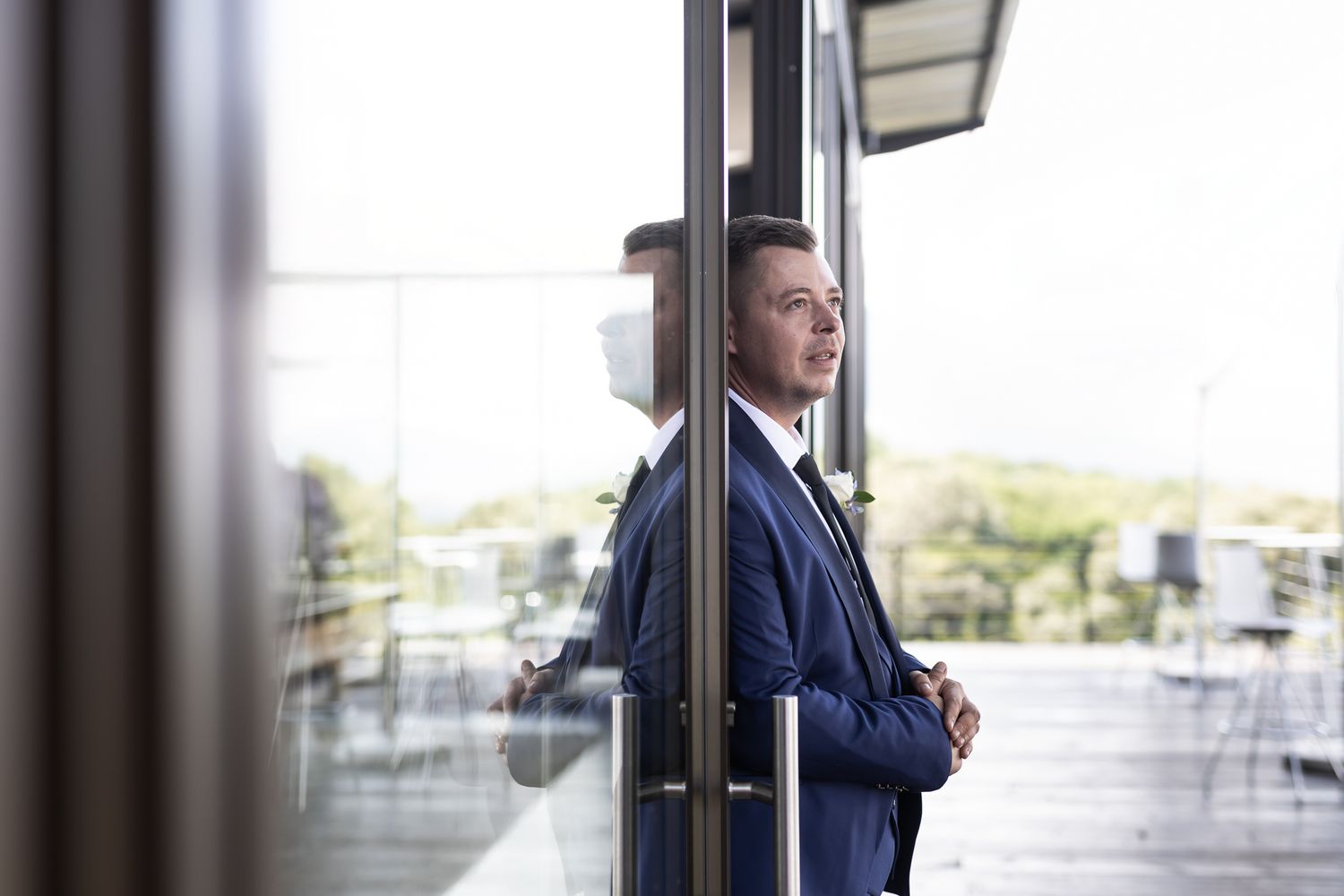 Person in navy suit gazes thoughtfully through glass doors on a balcony.
