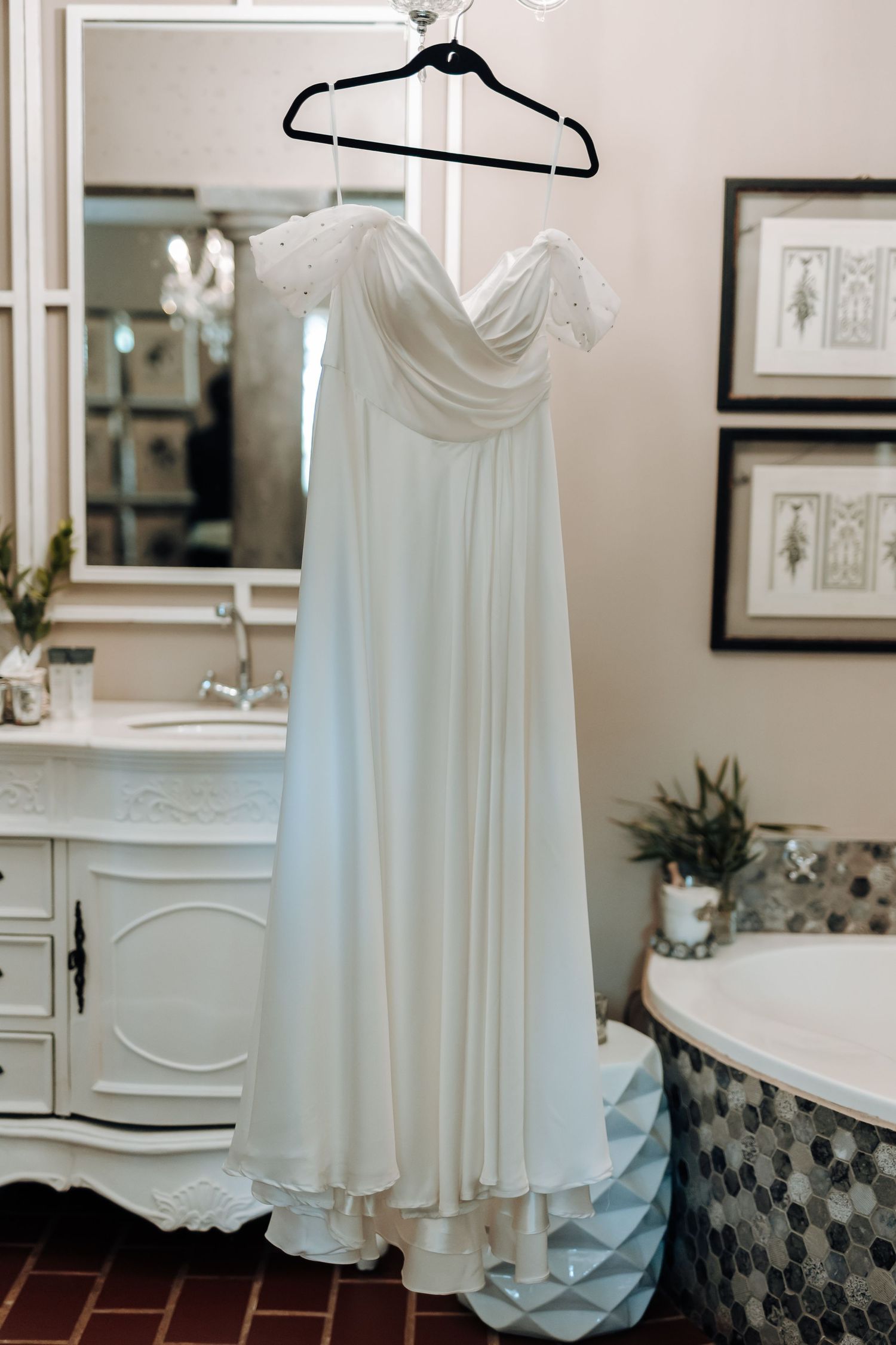 Flowing white wedding dress with off-shoulder design hanging on display in elegant dressing room.