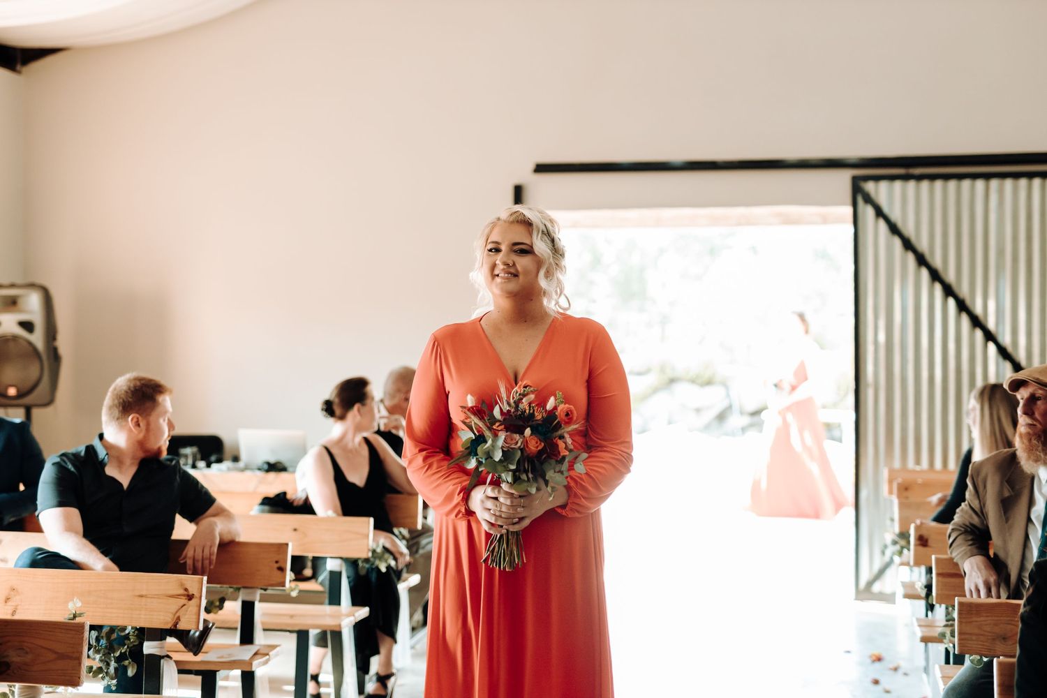 Bridesmaid in coral dress holds bouquet while walking down aisle at rustic indoor ceremony.