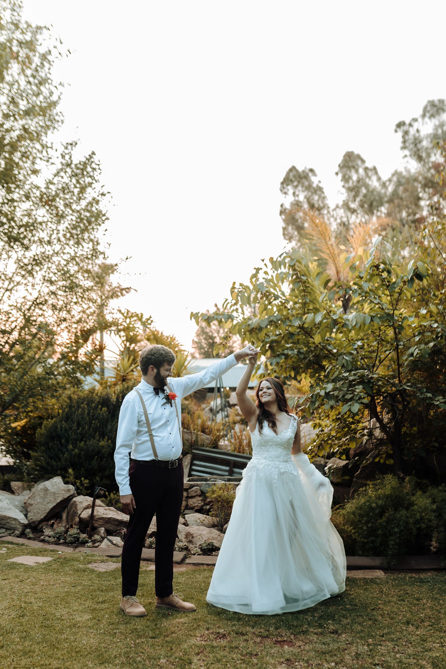 Wedding couple sharing joyful dance moment in sunlit garden setting.