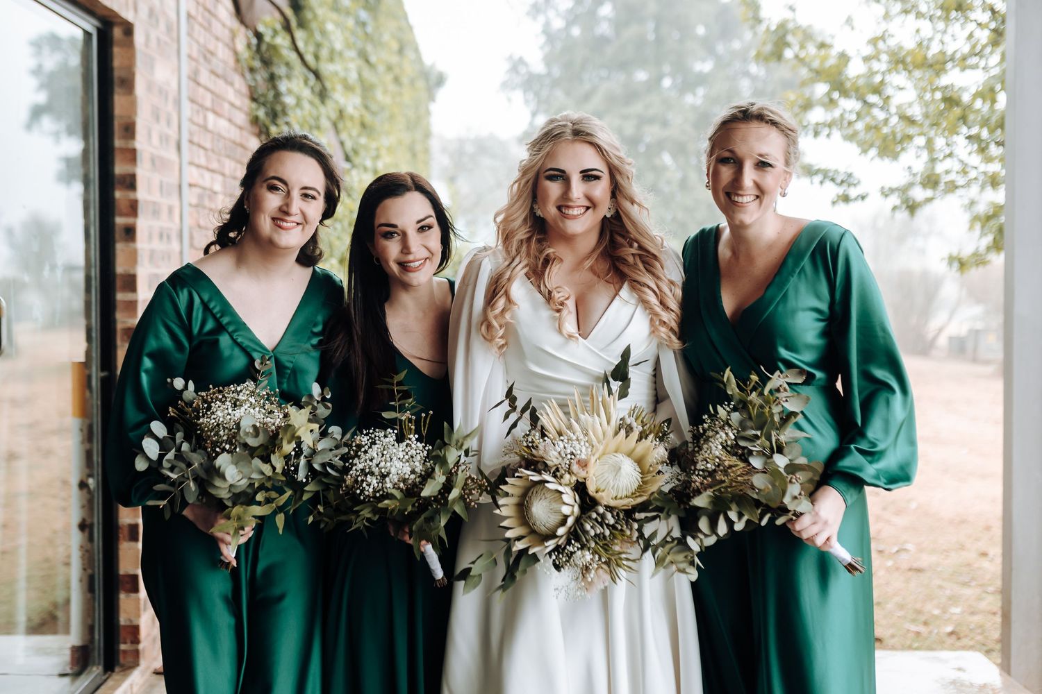 Bridal party in coordinating white and emerald dresses posing with dried flower bouquets.