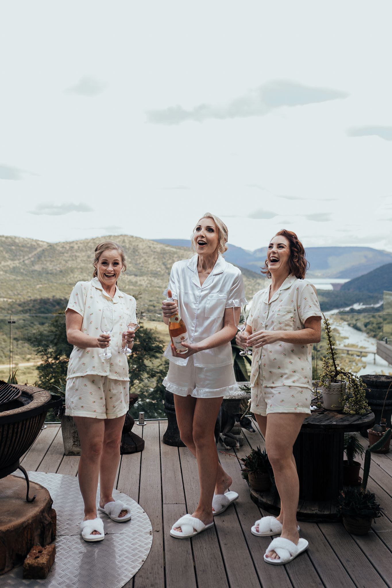 Friends laughing while celebrating in coordinated white outfits on scenic deck.